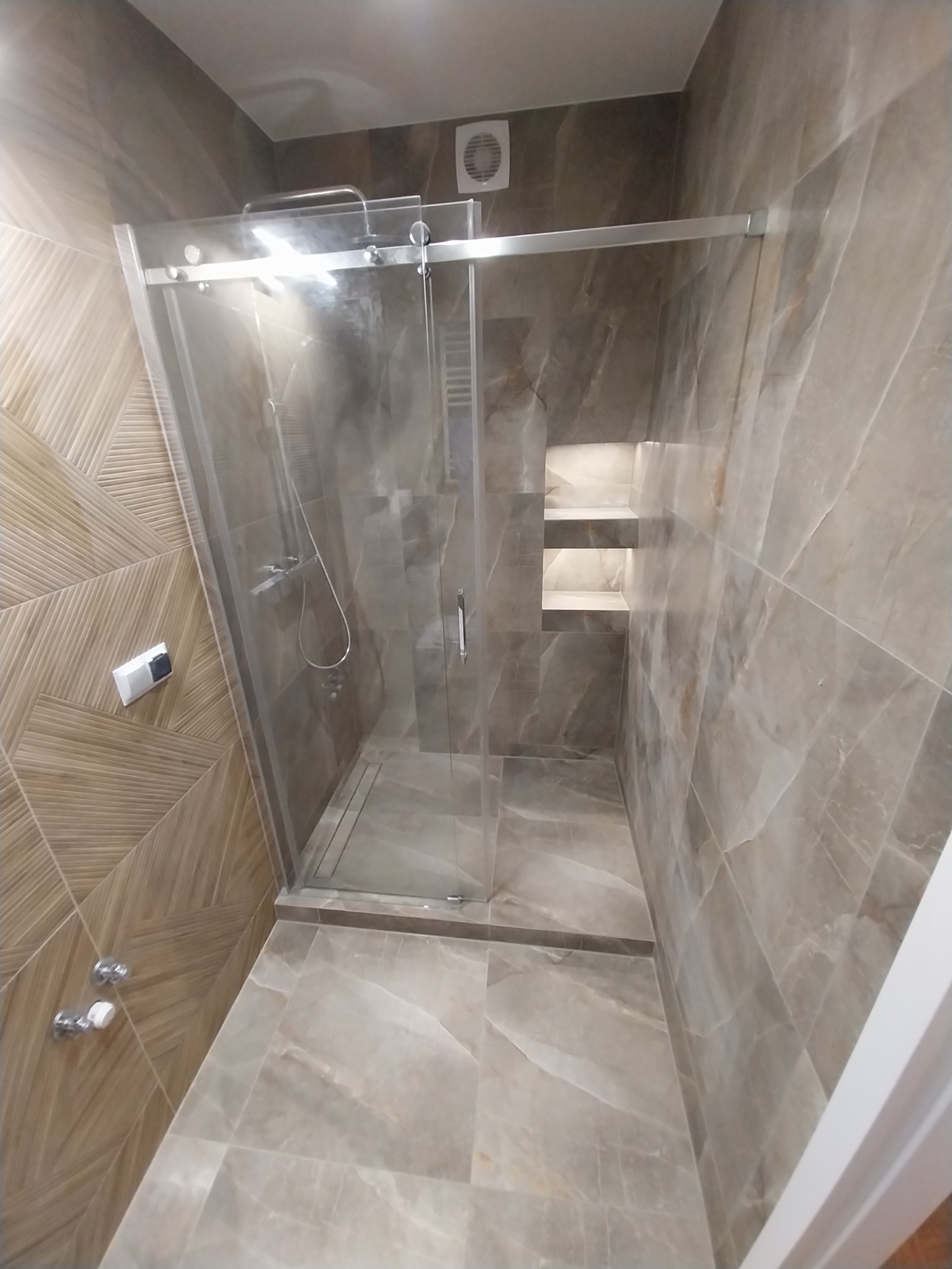Nowoczesna kabina prysznicowa z przesuwnymi drzwiami, wykończona płytkami imitującymi marmur i drewnem z geometrycznym wzorem na ścianie.