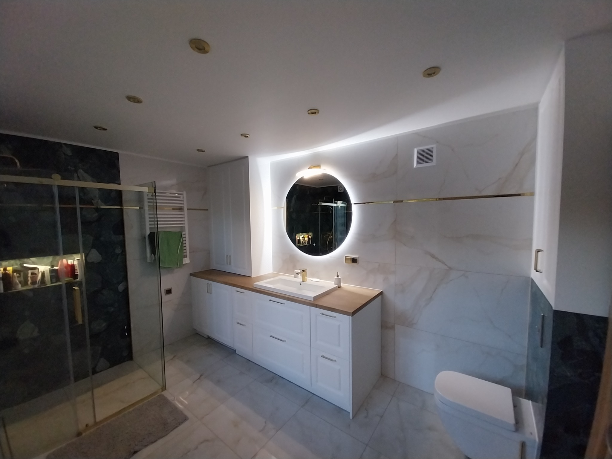 Nowoczesna łazienka z marmurowymi płytkami, okrągłym lustrem z podświetleniem LED, białymi szafkami pod umywalką z drewnianym blatem i kabiną prysznicową ze złotymi elementami.