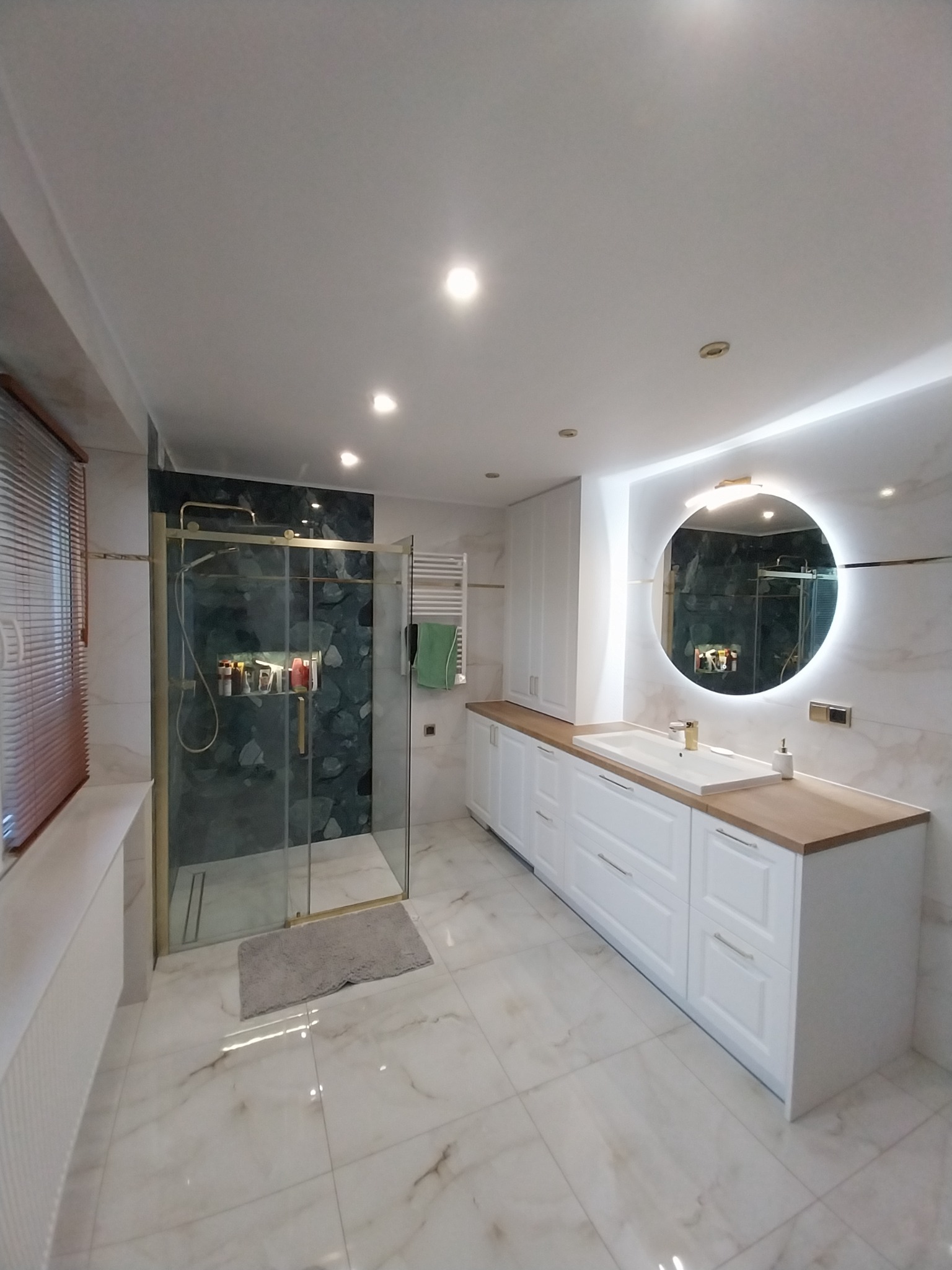 Nowoczesna łazienka z kabiną prysznicową ze złotymi ramami, okrągłym lustrem z podświetleniem LED, białą zabudową meblową z drewnianym blatem i marmurową posadzką.