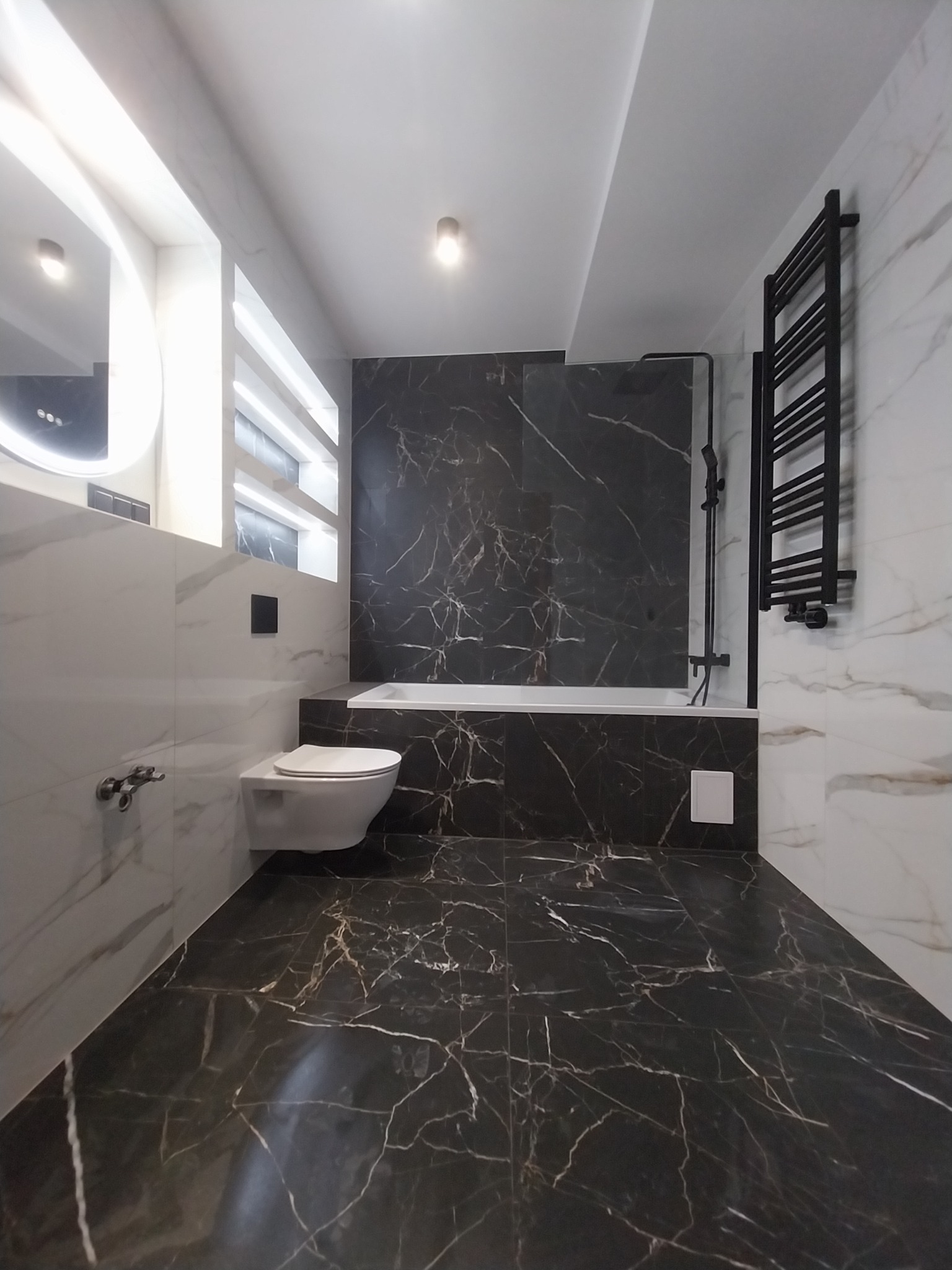 Elegancka łazienka w czarno-białej kolorystyce z wanną, podwieszaną toaletą, grzejnikiem drabinkowym i oświetleniem LED w niszach ściennych.