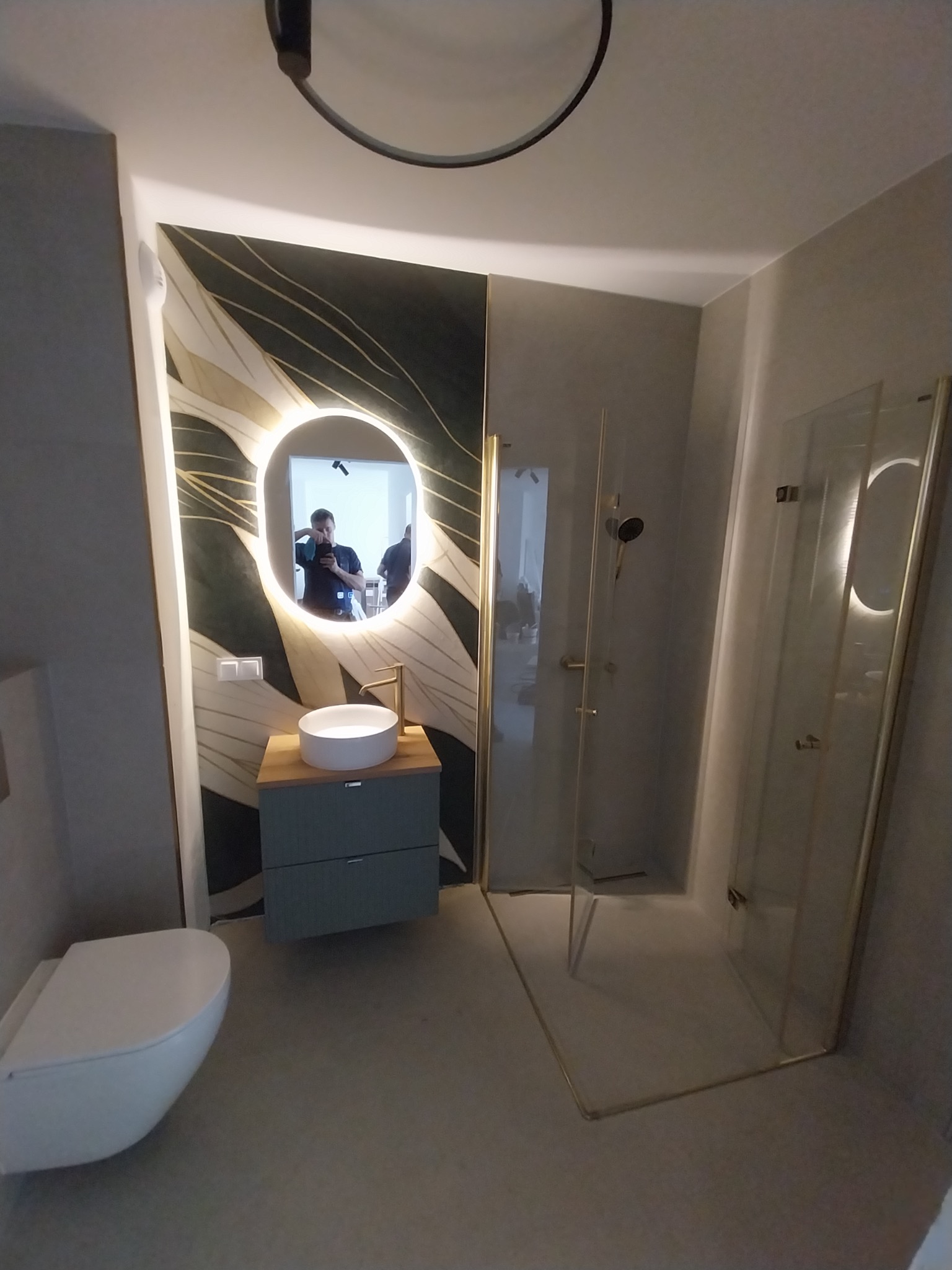 Nowoczesna łazienka z kabiną prysznicową ze złotymi ramami, umywalką na szafce i podświetlanym lustrem. Na ścianie dekoracyjny panel z motywem liści.