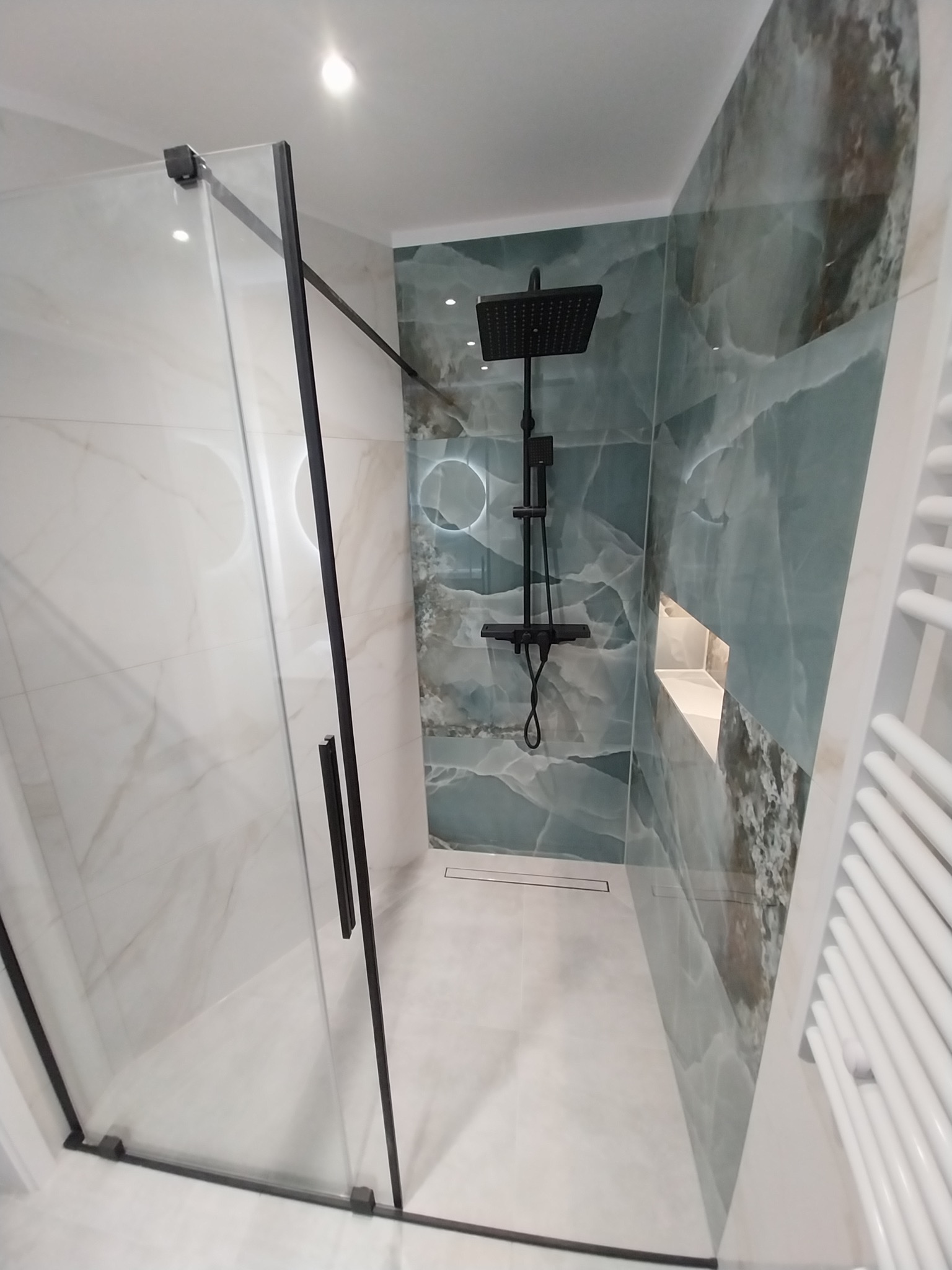 Nowoczesna kabina prysznicowa z czarną armaturą i płytkami imitującymi marmur w odcieniach bieli i turkusu. Widoczny odpływ liniowy i grzejnik drabinkowy.