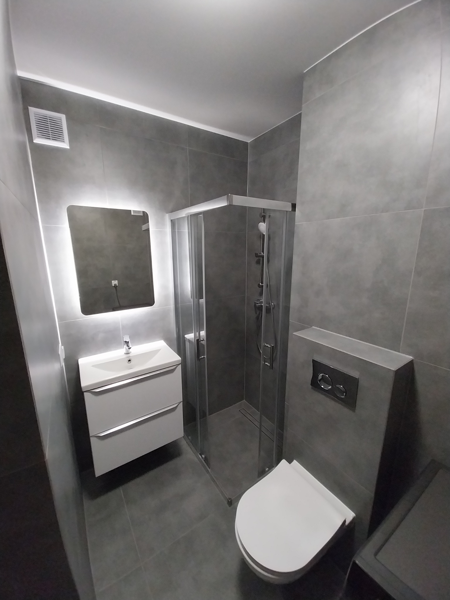 Nowoczesna, szara łazienka z kabiną prysznicową, podświetlanym lustrem nad białą szafką z umywalką i podwieszaną toaletą. Minimalistyczny design, płytki imitujące beton.