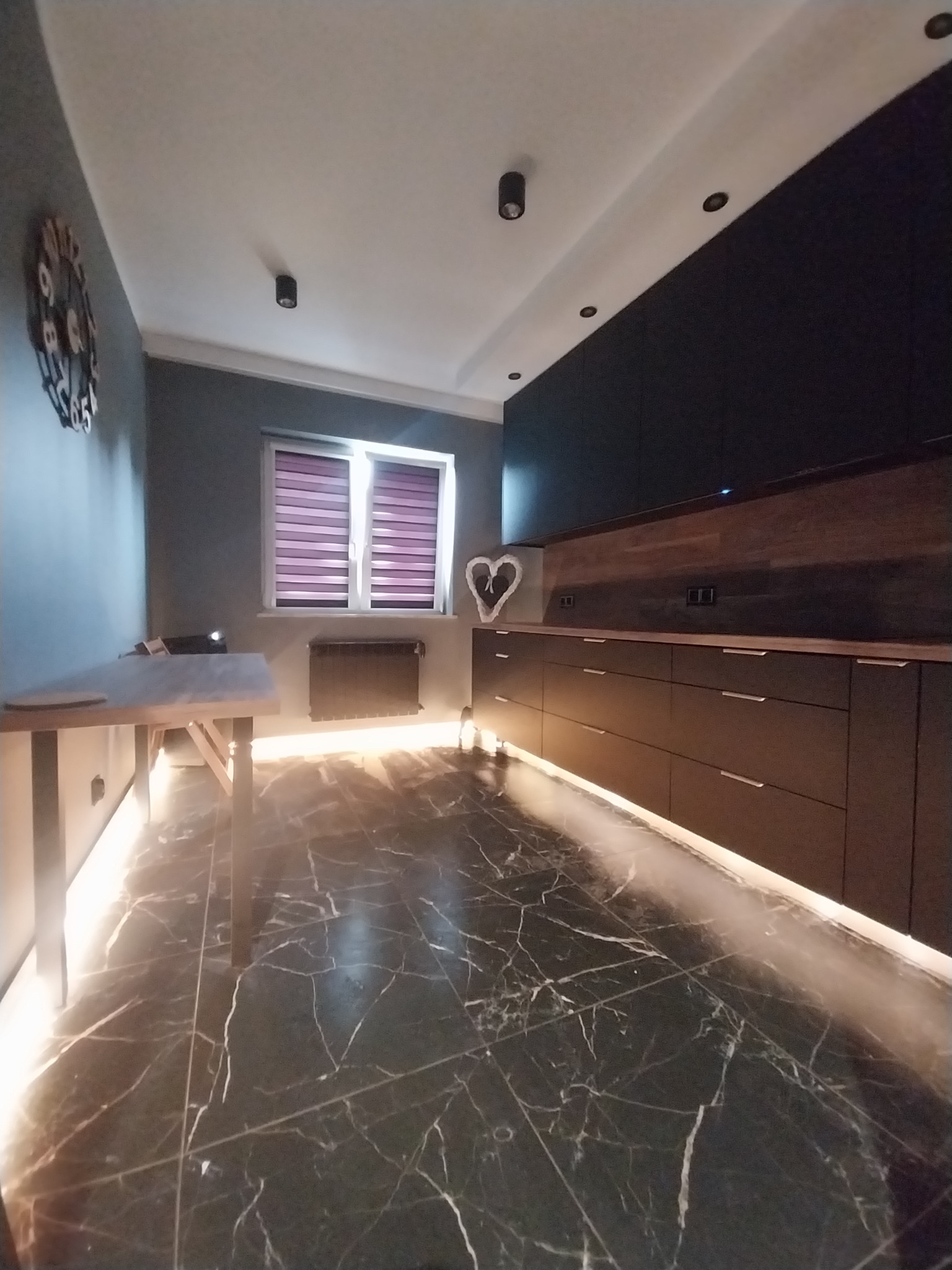 Nowoczesna kuchnia z czarnymi szafkami, marmurową podłogą i podświetleniem LED. Na ścianie zegar i dekoracja w kształcie serca. Okno z żaluzjami.