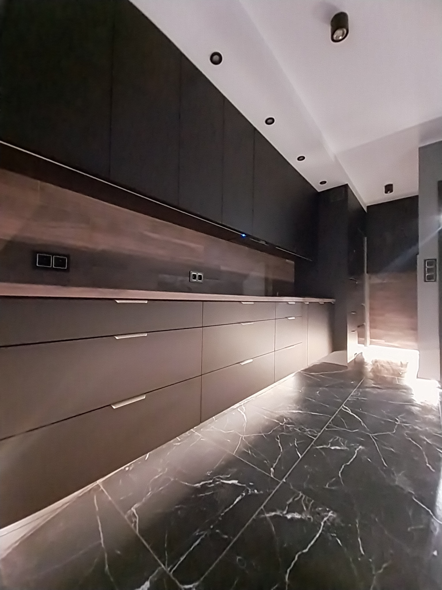 Nowoczesna łazienka z ciemnymi, matowymi szafkami z podłużnymi uchwytami i marmurową podłogą w odcieniach szarości i bieli. Oświetlenie punktowe sufitowe.