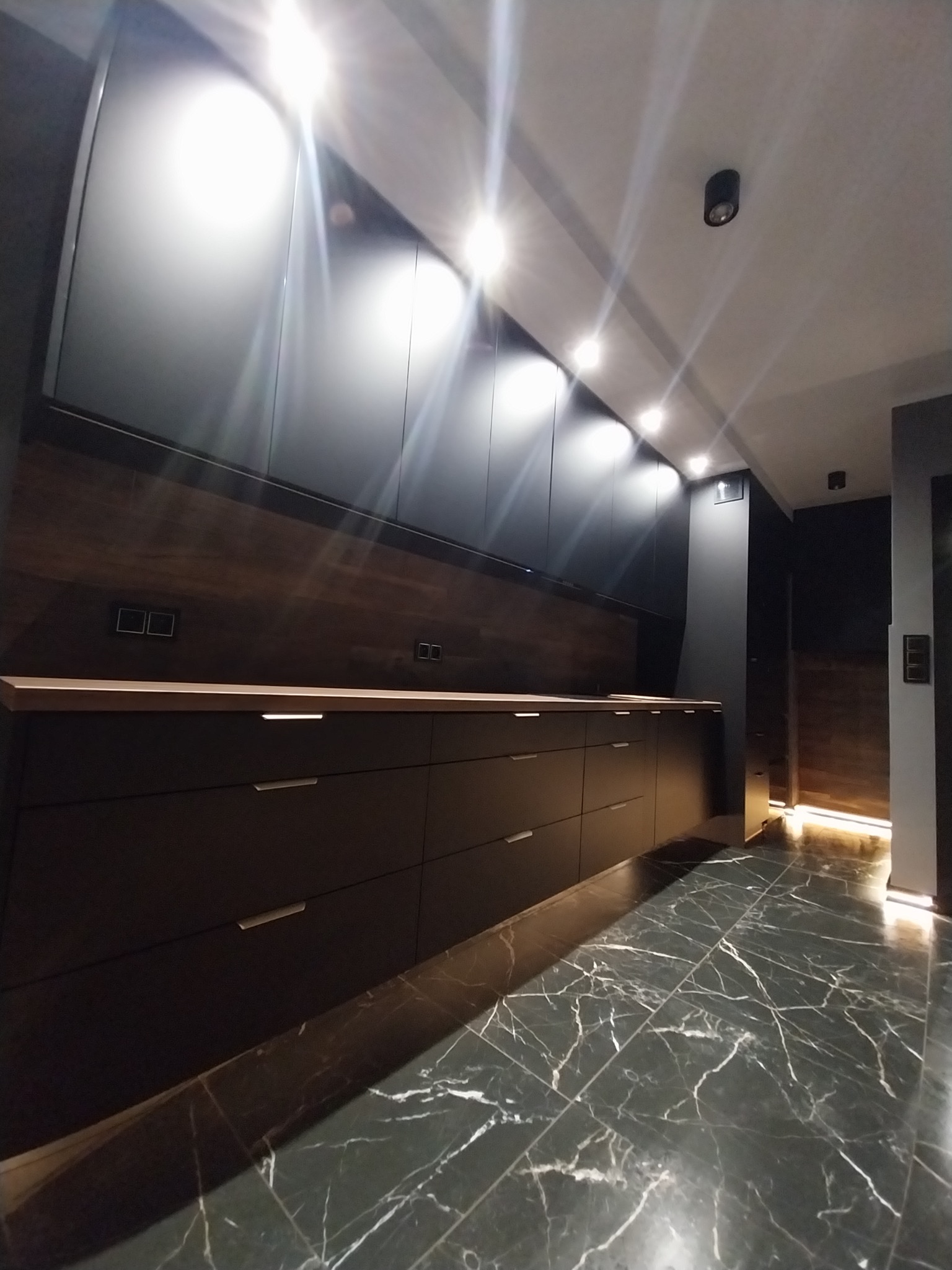 Nowoczesna, ciemna kuchnia z marmurową podłogą i oświetleniem punktowym. Długi blat z drewnianym akcentem i minimalistyczne szafki tworzą elegancką przestrzeń.