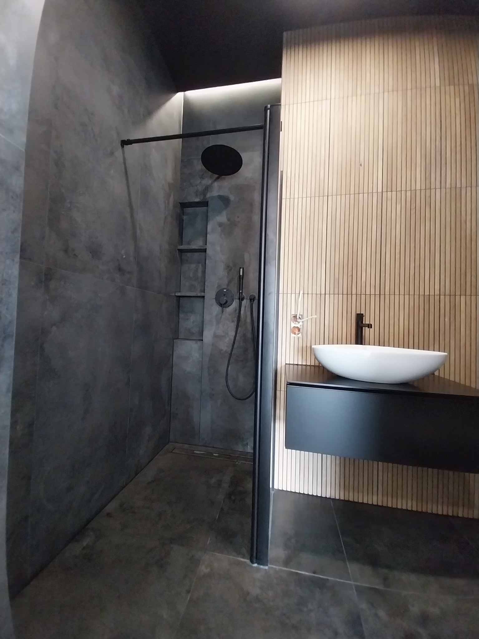 Nowoczesna łazienka z kabiną prysznicową typu walk-in, szare ściany z wnękami i drewnopodobne panele. Umywalka nablatowa na czarnej szafce podwieszanej.