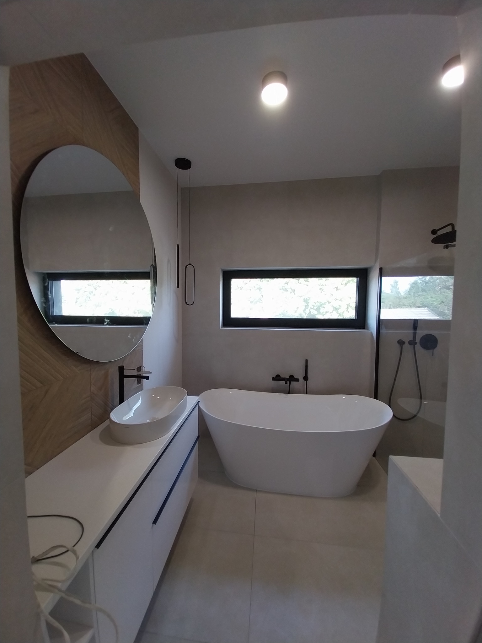 Nowoczesna łazienka z owalnym lustrem, białą wolnostojącą wanną i minimalistyczną armaturą w czarnym kolorze. Ściana z drewnianą okładziną w jodełkę.