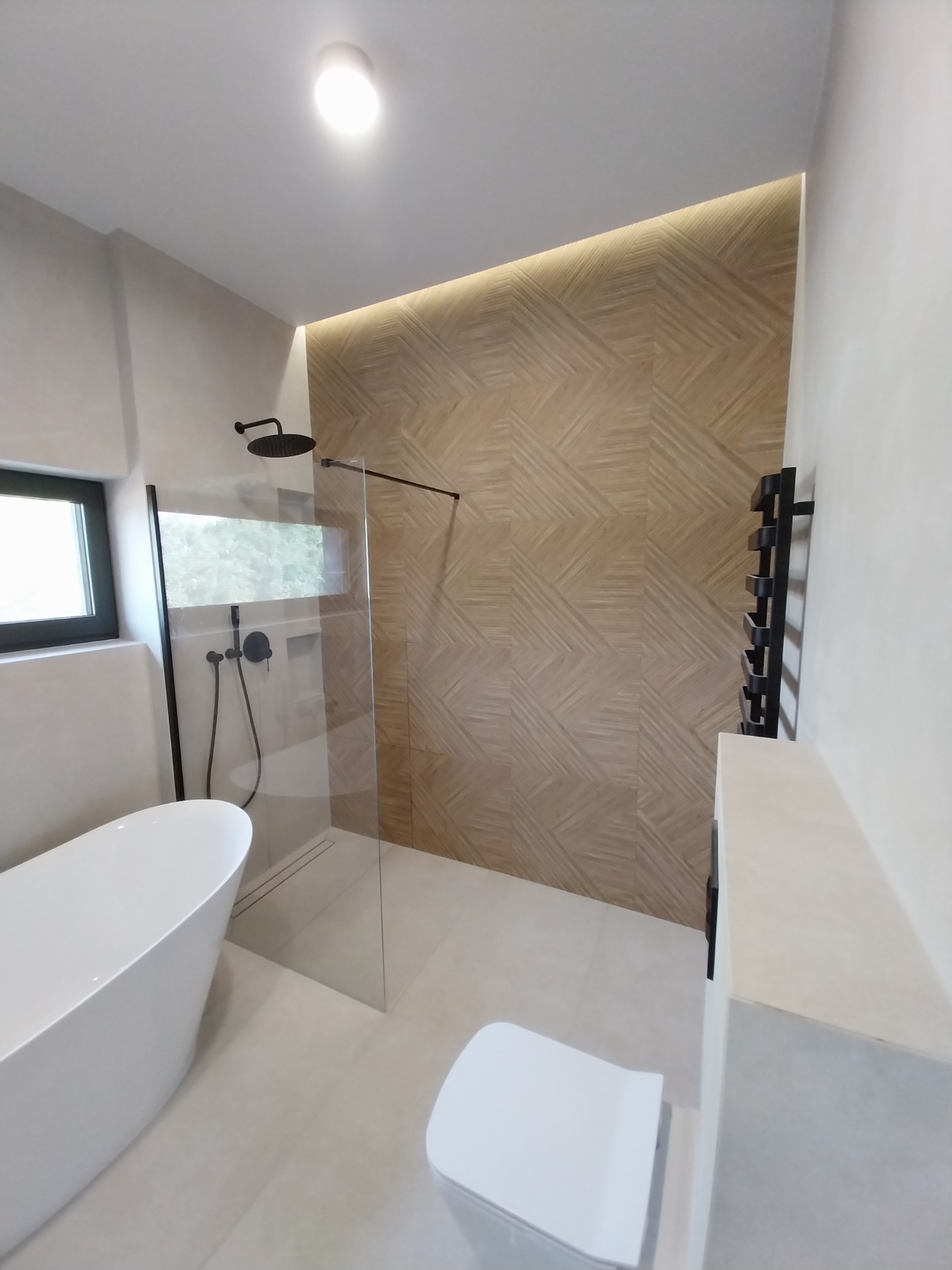 Nowoczesna łazienka z wolnostojącą wanną, kabiną prysznicową typu walk-in i ścianą z drewnopodobnych płytek z podświetleniem LED. Minimalistyczny design, czarne akcenty.