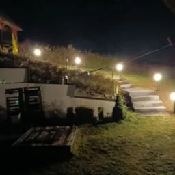 Oświetlony ogród nocą z betonowymi schodami prowadzącymi w górę, lampy kuliste wzdłuż ścieżki, drewniana altana w tle, widoczne elementy małej architektury.