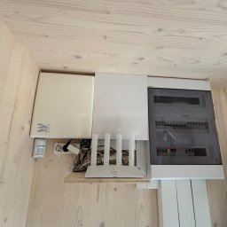 KAPI Kacper Dułak - Biała skrzynka elektryczna z bezpiecznikami, router WiFi i czujnik ruchu na drewnianej ścianie. Widoczne kable i przewody. Ujęcie z dołu, oświetlenie naturalne.