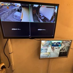 KAPI Kacper Dułak - Dwa monitory CCTV zamontowane na ścianie, prezentujące obraz z kamer monitoringu. Na górnym monitorze widok z czterech kamer, na dolnym z ośmiu.