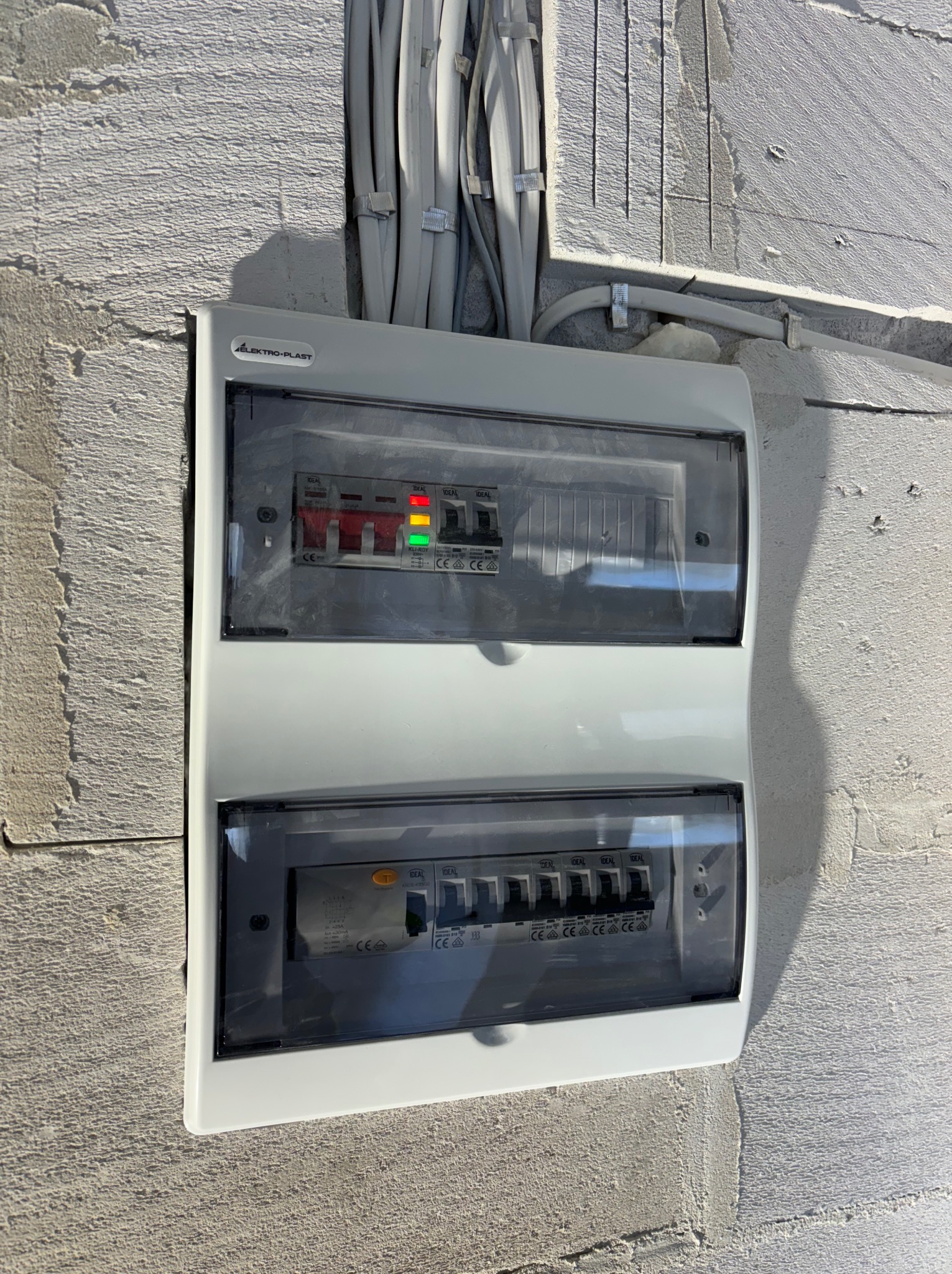 Szara skrzynka rozdzielcza z bezpiecznikami, zamontowana na surowej, szarej ścianie. Widoczne kable prowadzone w listwach mocowanych do ściany.