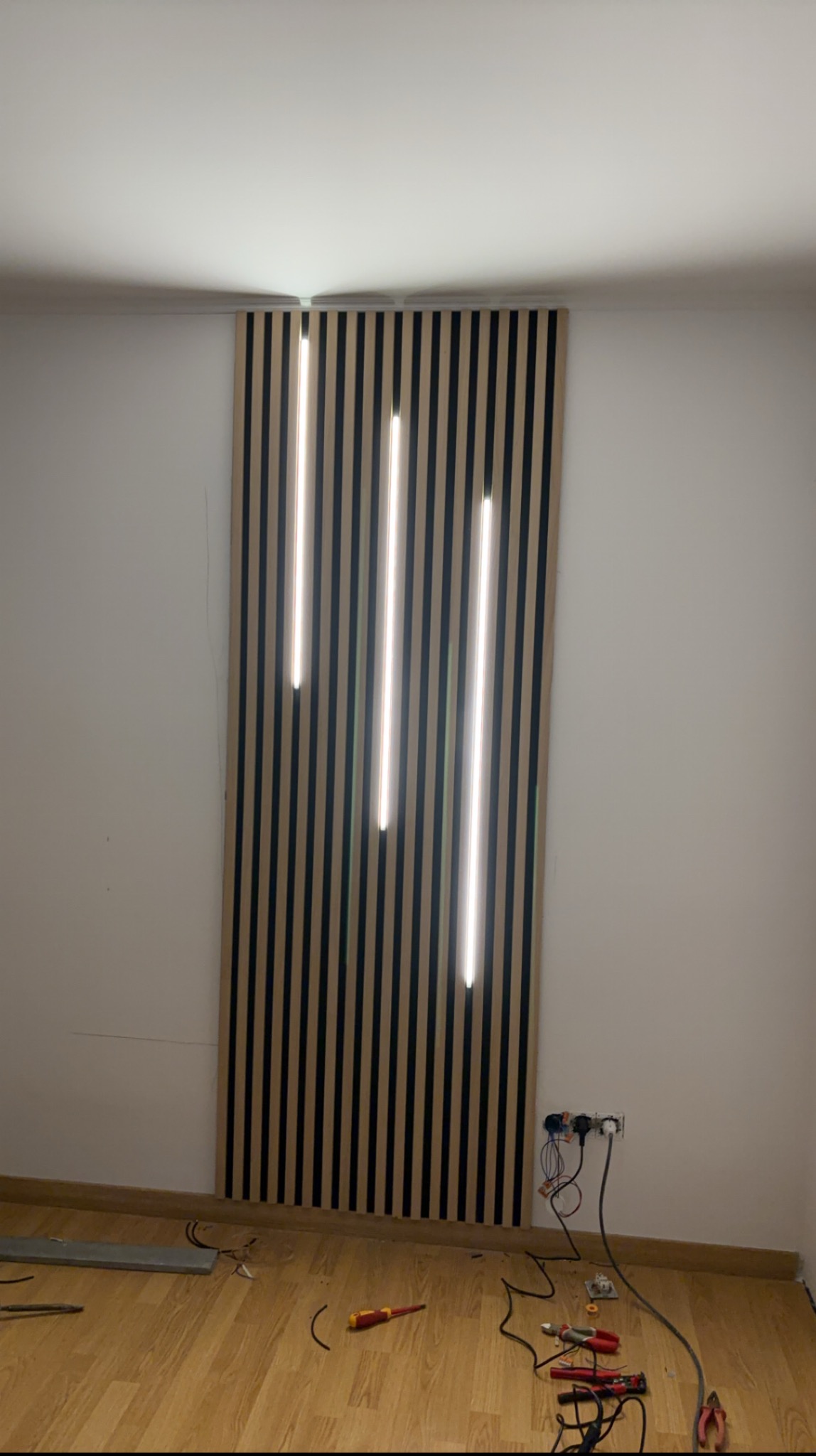 Panel dekoracyjny z pionowymi lamelami drewnianymi i trzema wbudowanymi pionowymi lampami LED, widoczne kable i narzędzia na podłodze podczas montażu.