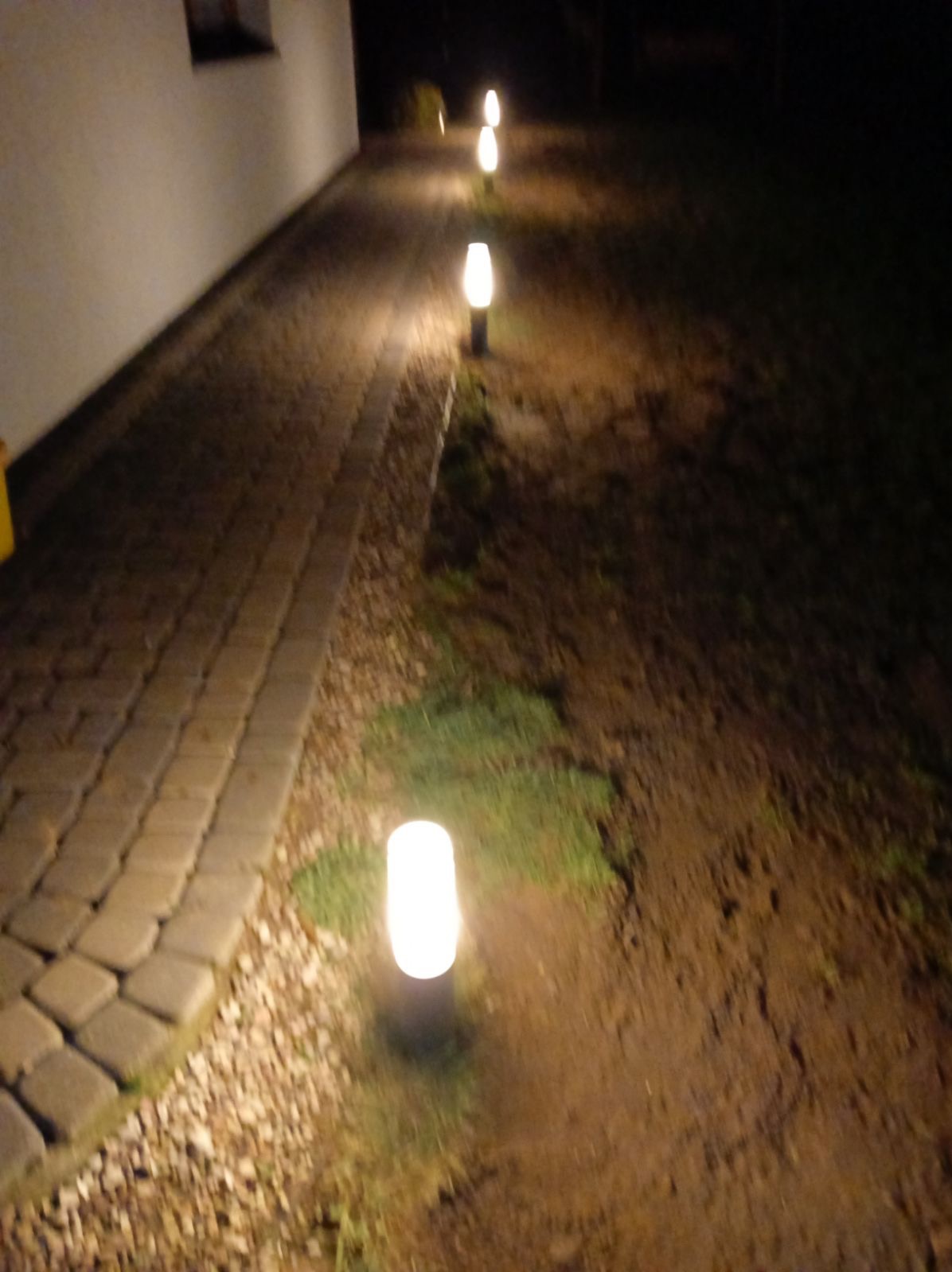 Ścieżka oświetlona szeregiem lamp ogrodowych w kształcie walców, widoczna nocą, z bliska ścieżka z kostki brukowej.