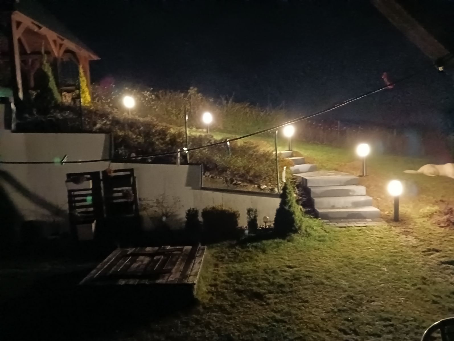 Oświetlony ogród nocą z betonowymi schodami prowadzącymi w górę, lampy kuliste wzdłuż ścieżki, drewniana altana w tle, widoczne elementy małej architektury.