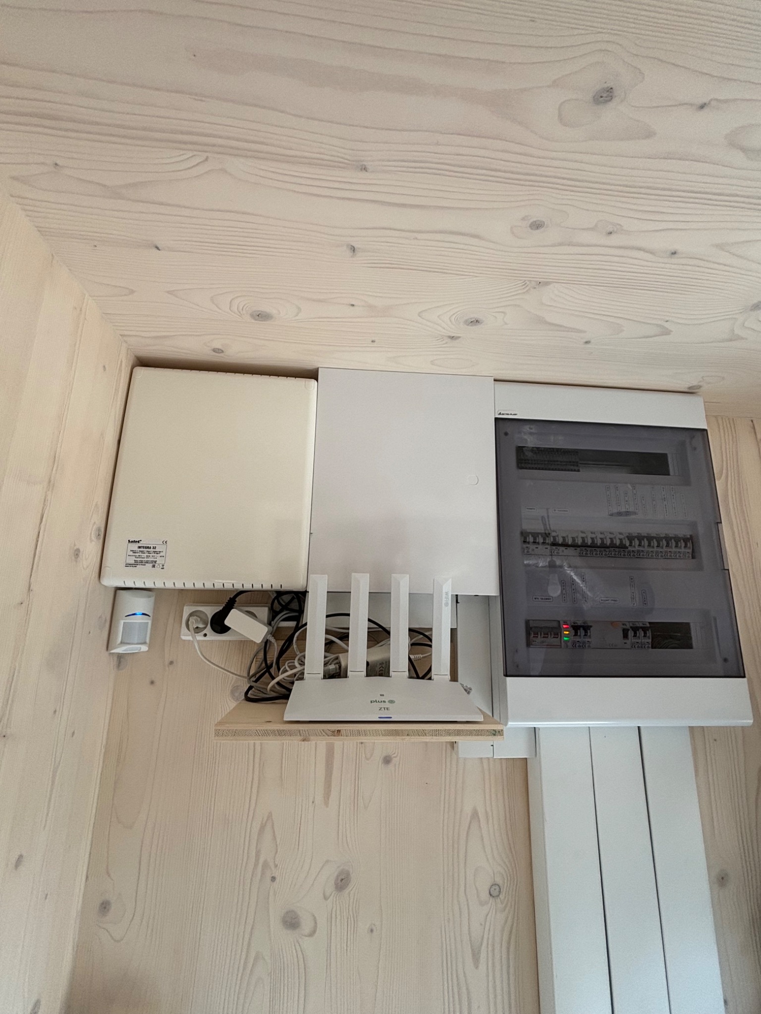 Biała skrzynka elektryczna z bezpiecznikami, router WiFi i czujnik ruchu na drewnianej ścianie. Widoczne kable i przewody. Ujęcie z dołu, oświetlenie naturalne.