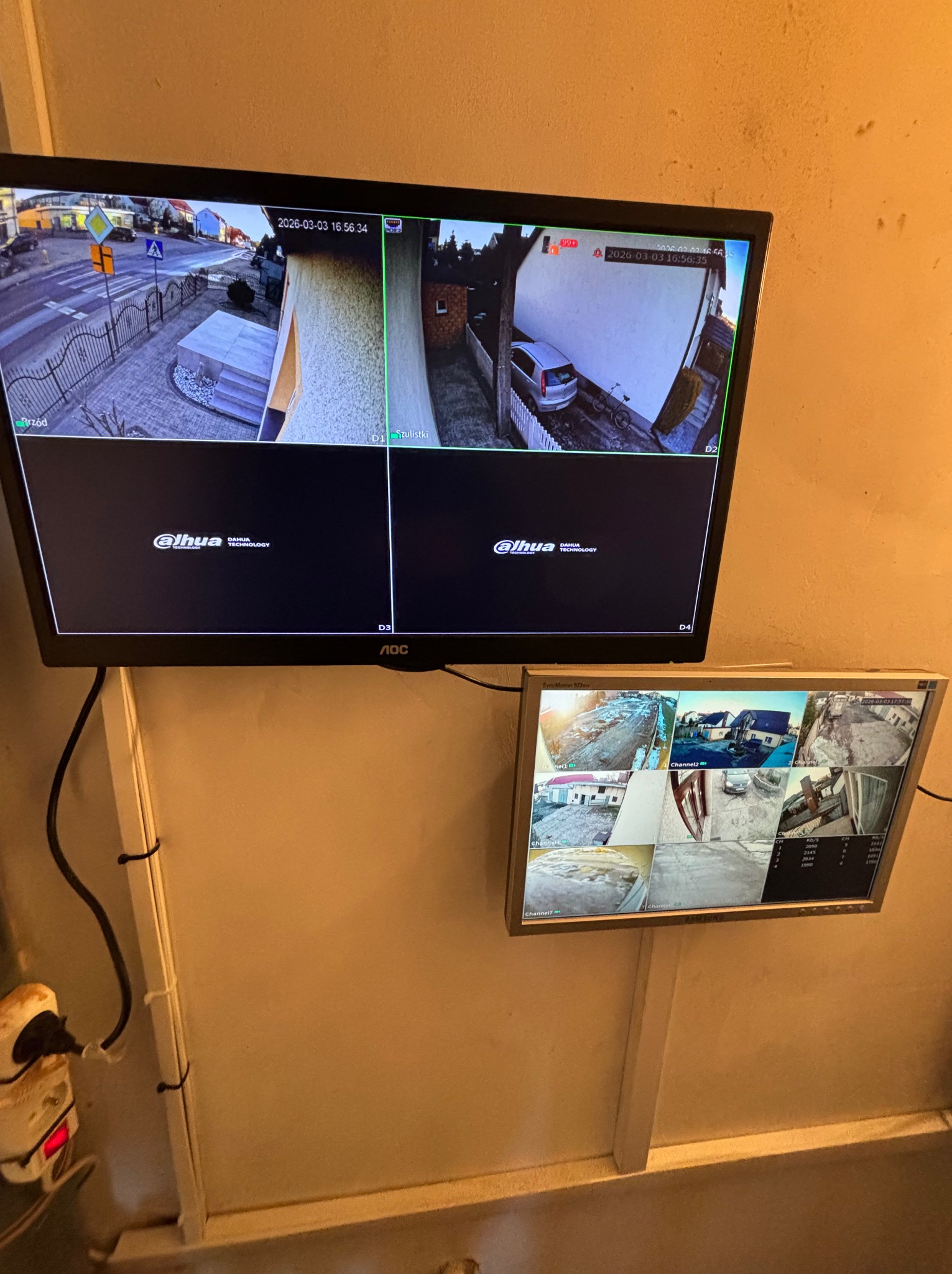 Dwa monitory CCTV zamontowane na ścianie, prezentujące obraz z kamer monitoringu. Na górnym monitorze widok z czterech kamer, na dolnym z ośmiu.