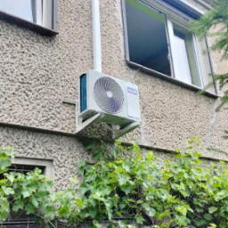 Zewnętrzna jednostka klimatyzatora marki AUX zamontowana na ścianie budynku z oknami, z widoczną instalacją odprowadzającą skropliny i pnączami winorośli w dolnej części kadru.