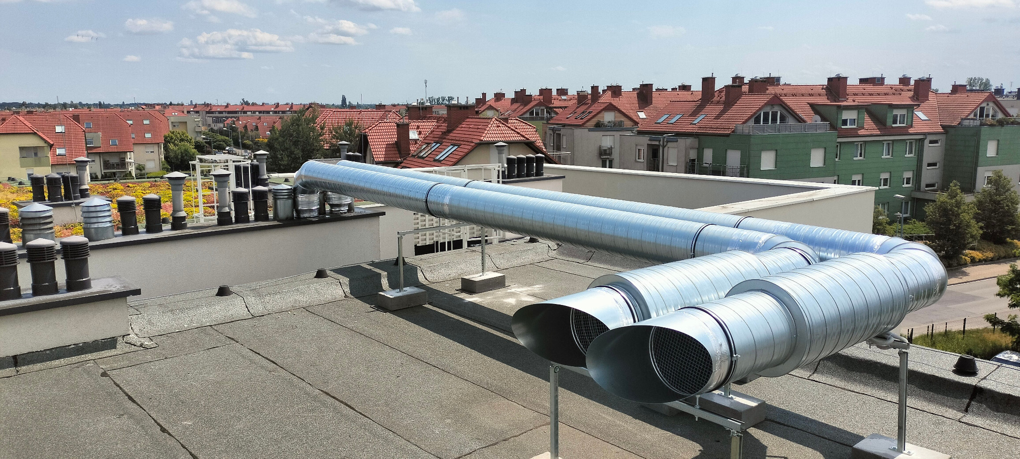 Dwa równoległe, spiralnie zwijane, metalowe przewody wentylacyjne na dachu budynku z widokiem na osiedle domów jednorodzinnych z czerwonymi dachami.