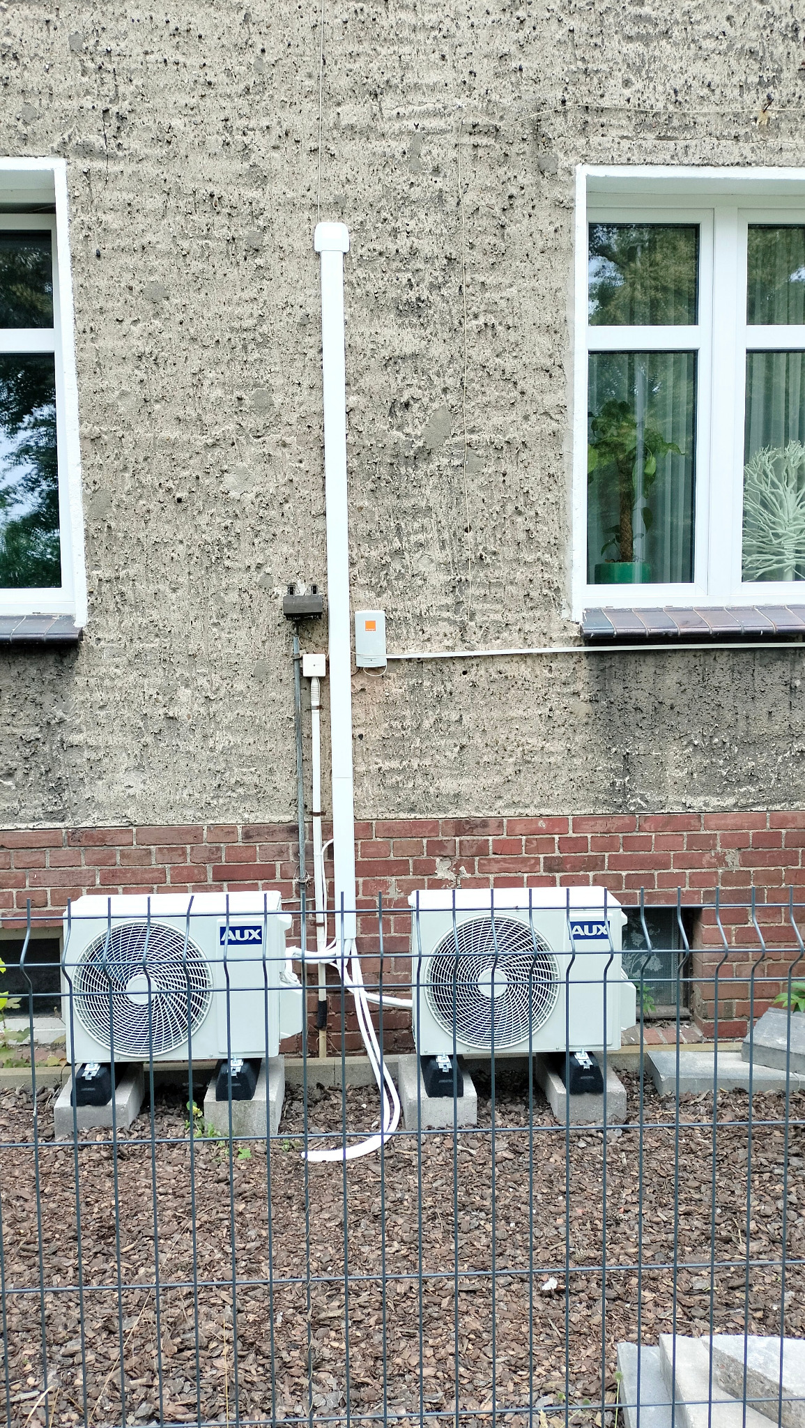 Dwa identyczne, jasnoszare agregaty klimatyzacyjne marki AUX, zamontowane na betonowych podstawach przed budynkiem z oknami i ścianą z cegły, widoczne przez metalowe ogrodzenie.