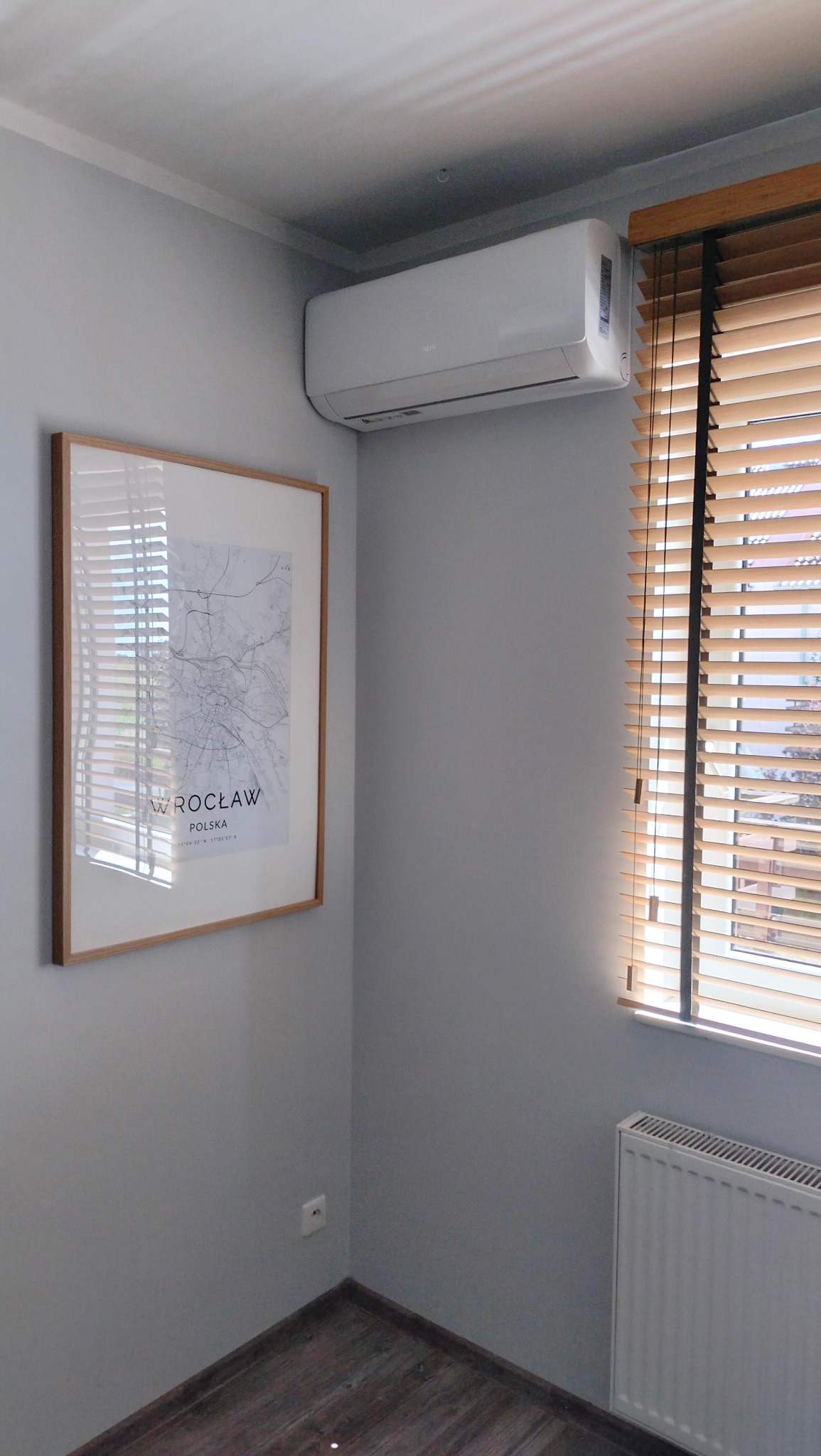 Biały klimatyzator ścienny zamontowany w szarym pokoju obok okna z drewnianymi żaluzjami i obrazu z mapą Wrocławia w ramce.