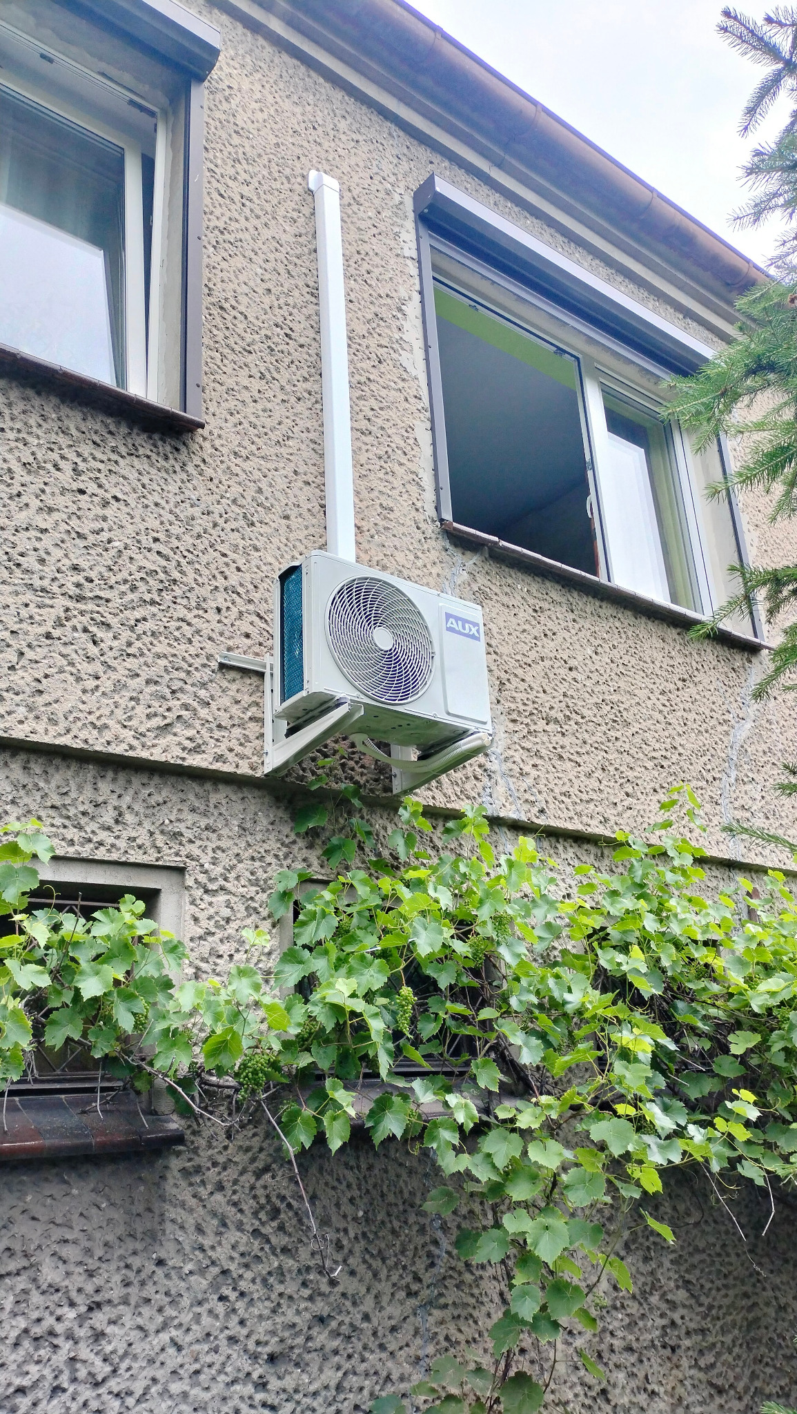 Zewnętrzna jednostka klimatyzatora marki AUX zamontowana na ścianie budynku z oknami, z widoczną instalacją odprowadzającą skropliny i pnączami winorośli w dolnej części kadru.