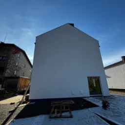 Nowo otynkowana ściana szczytowa domu jednorodzinnego w słoneczny dzień, z widocznymi elementami zabezpieczającymi teren budowy, takimi jak folia ochronna i paleta.