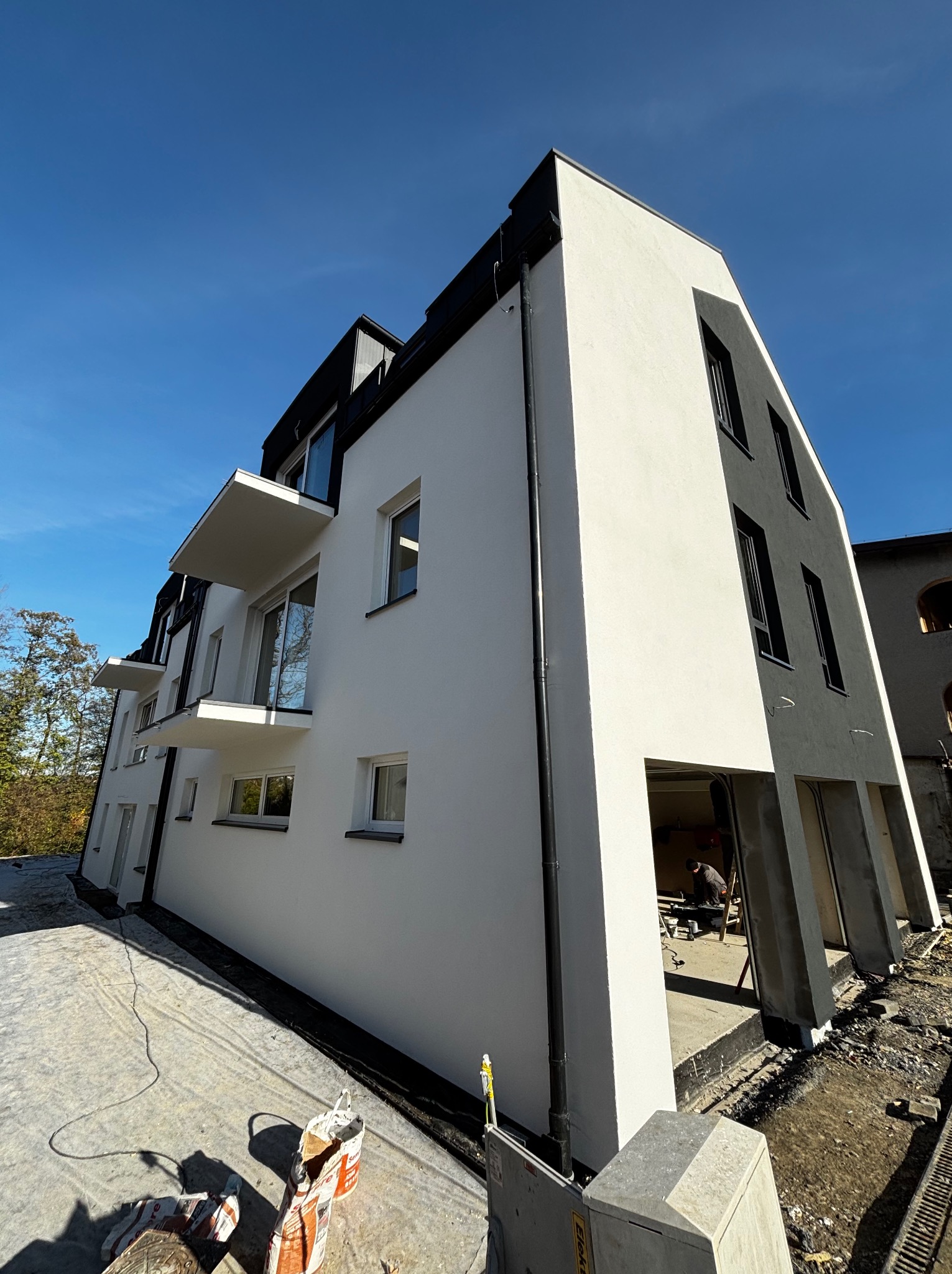 Nowoczesny, dwukondygnacyjny budynek z balkonami, świeżo otynkowany na biało, z czarnymi detalami architektonicznymi, widok z dołu w słoneczny dzień, teren wokół budynku w trakcie prac...