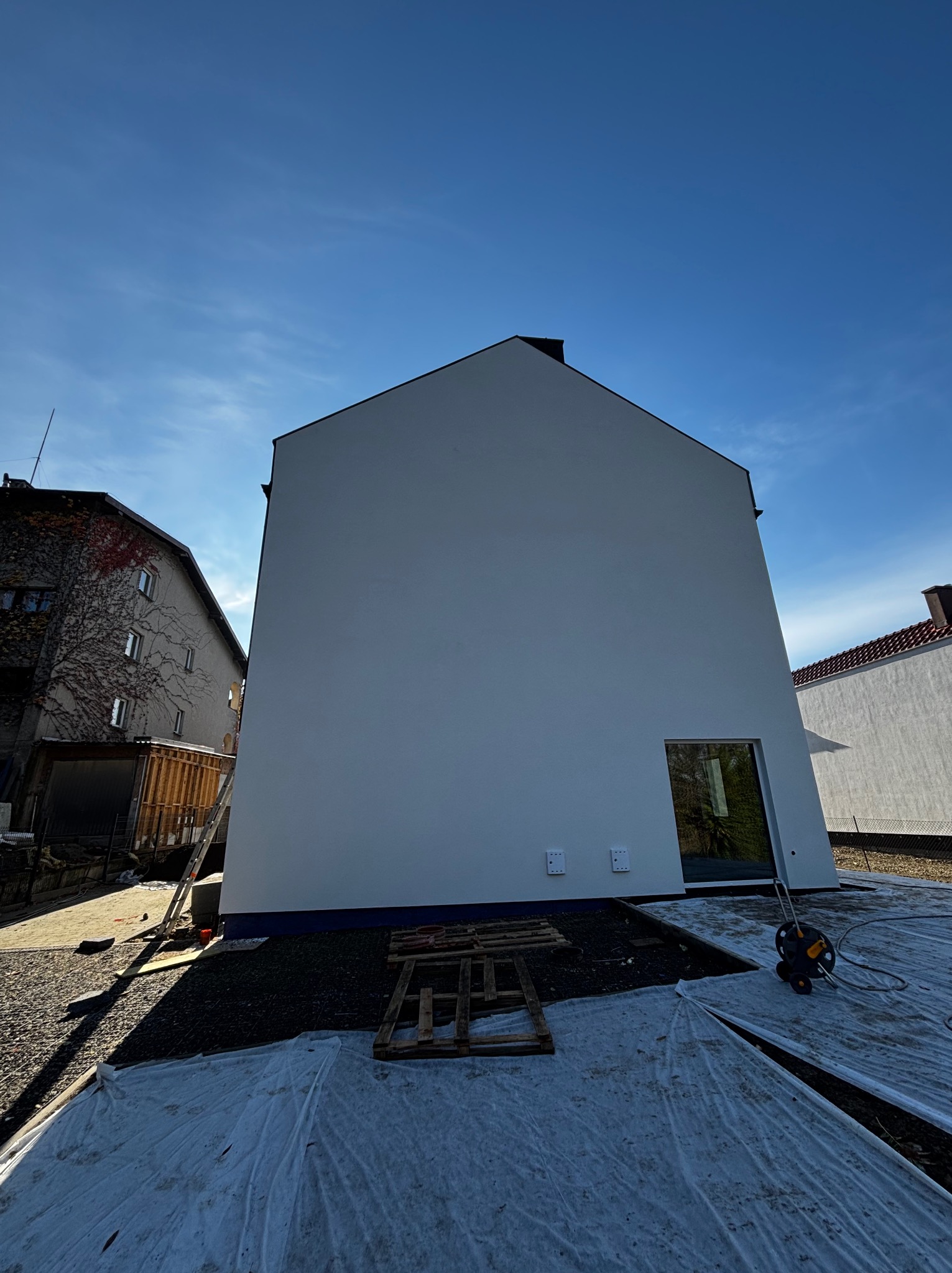 Nowo otynkowana ściana szczytowa domu jednorodzinnego w słoneczny dzień, z widocznymi elementami zabezpieczającymi teren budowy, takimi jak folia ochronna i paleta.