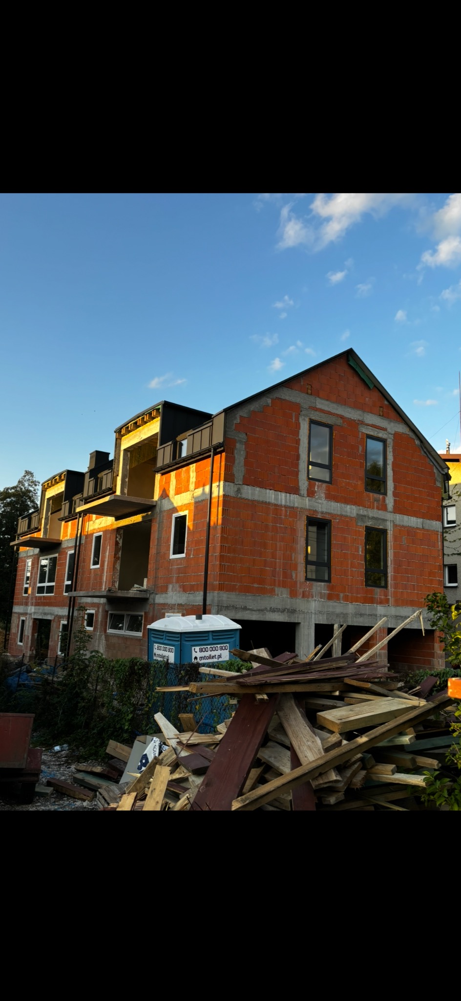Surowy stan budynku wielorodzinnego z czerwonej cegły w trakcie budowy, widoczne niezamontowane okna, na pierwszym planie sterta drewnianych odpadów i toaleta przenośna.