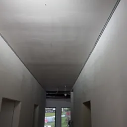 Korytarz w trakcie wykańczania: gładkie, białe ściany i sufit z widocznymi listwami sufitowymi, narzędzia malarskie w tle, widok na zewnątrz przez okno.