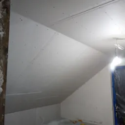 Poddasze w trakcie wykańczania: ściany i skośny sufit pokryte płytami gipsowo-kartonowymi, widoczne łączenia i ślady po wkrętach, narzędzia i resztki materiałów na podłodze.