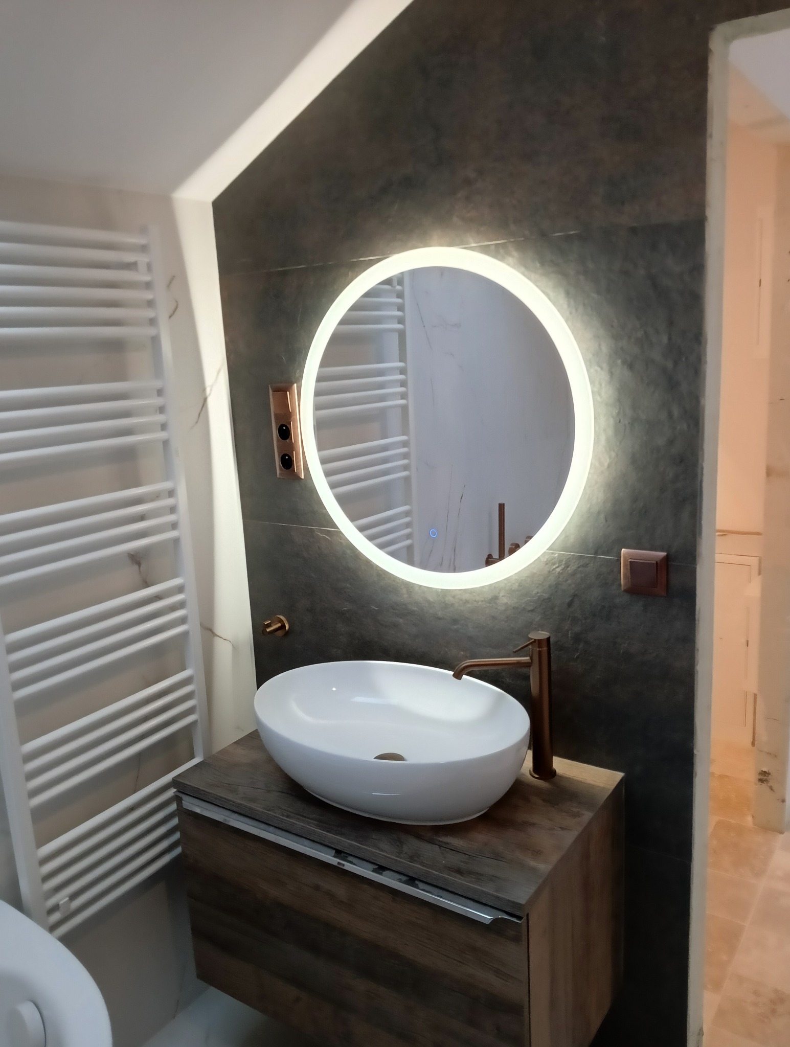 Nowoczesna łazienka z okrągłym lustrem LED, umywalką nablatową i grzejnikiem drabinkowym. Ciemna ściana z elementami miedzianymi.