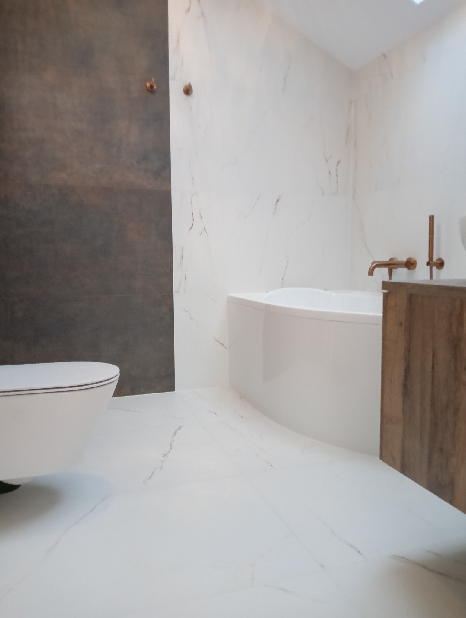 Nowoczesna łazienka z białymi marmurowymi płytkami, brązową ścianą i armaturą w kolorze miedzi. Uwagę przykuwa minimalistyczna toaleta i wanna.