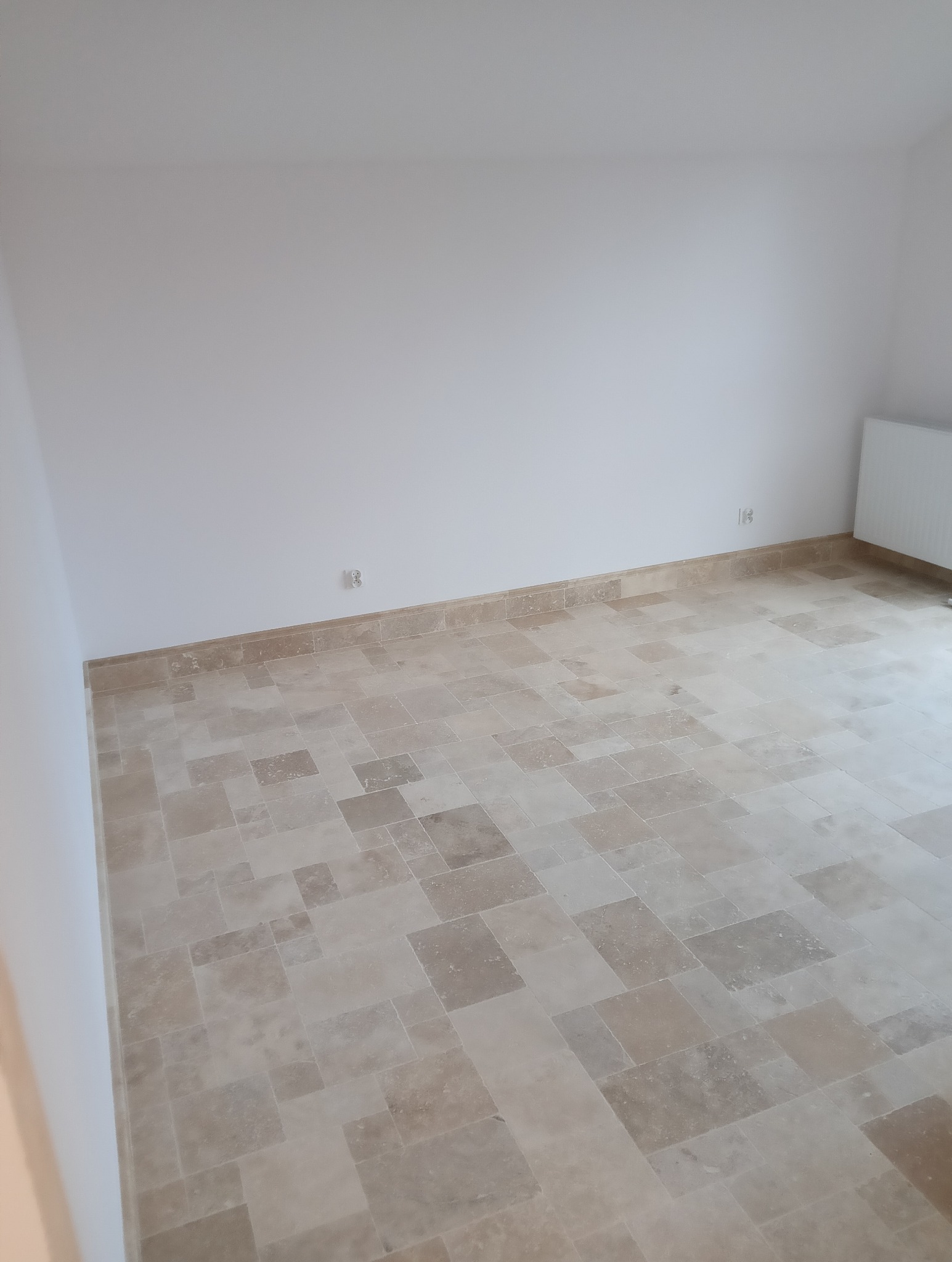 Wykończone wnętrze z beżową, trawertynową podłogą ułożoną w nieregularny wzór. Białe ściany, widoczne gniazdka elektryczne i grzejnik. Minimalistyczny styl.