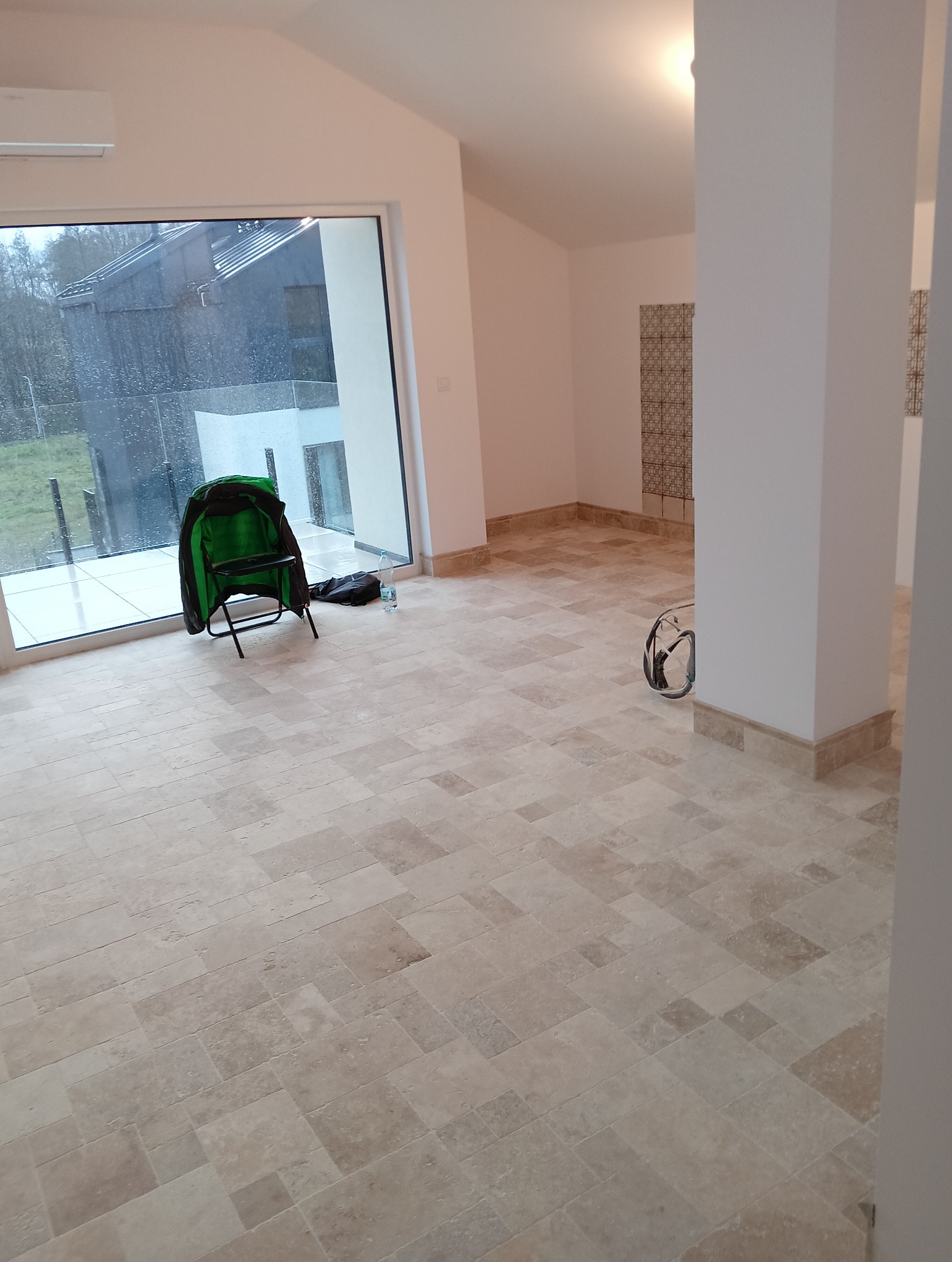 Jasna, beżowa podłoga z płytek w nowym wnętrzu. Widoczne duże okno z widokiem na zewnątrz, krzesło turystyczne i butelka wody. Wykończenie podłogi z listwami przypodłogowymi.