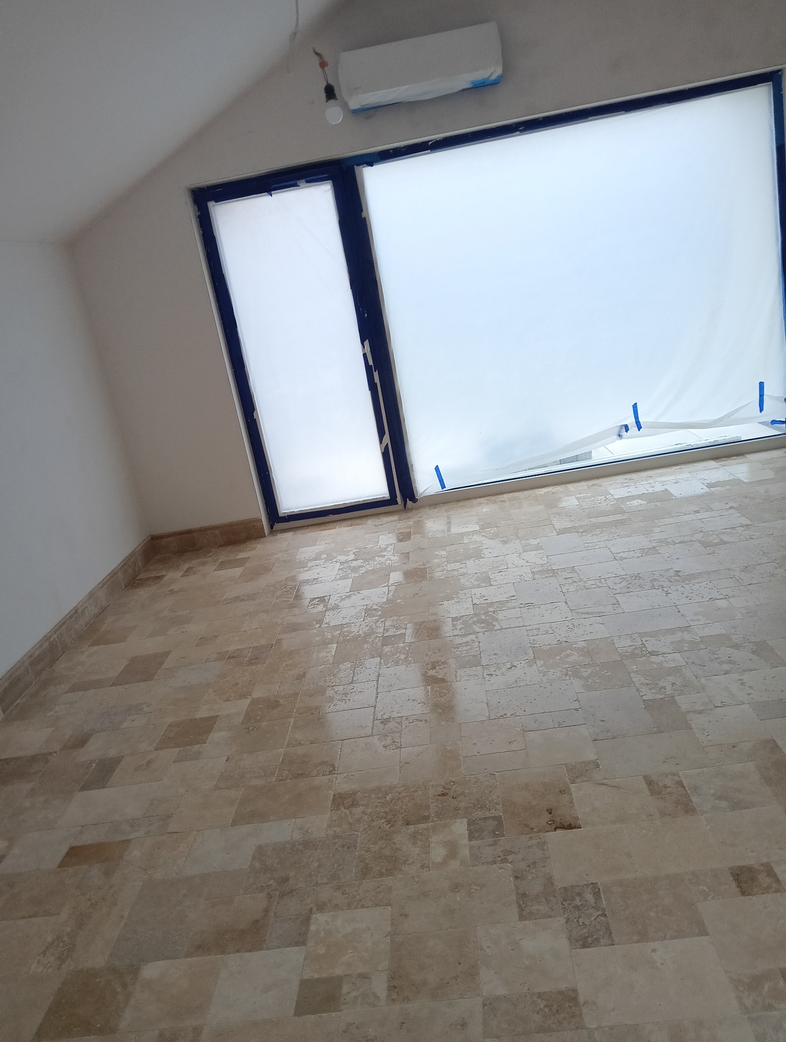 Jasna, nowa podłoga z płytek w pokoju z oknem zabezpieczonym folią malarską. Widoczny klimatyzator i sufit ze skosem. Ukończone prace glazurnicze.