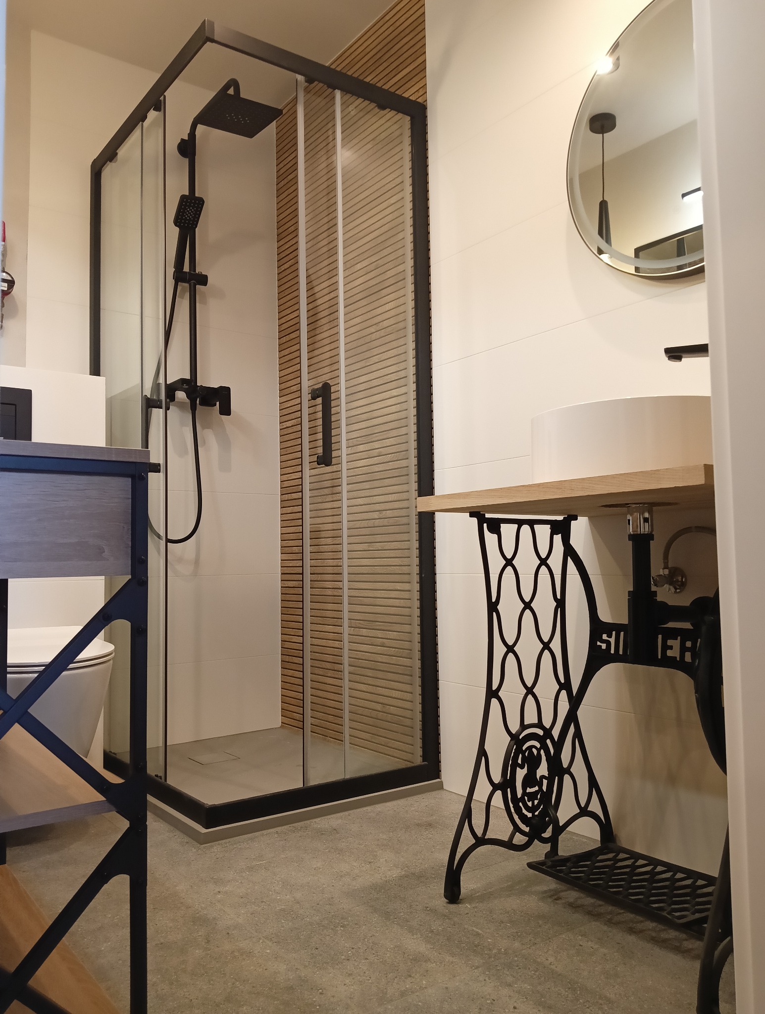 Nowoczesna łazienka z kabiną prysznicową typu walk-in z czarnymi ramami, umywalką na blacie z podstawą maszyny do szycia Singer i okrągłym lustrem z podświetleniem.