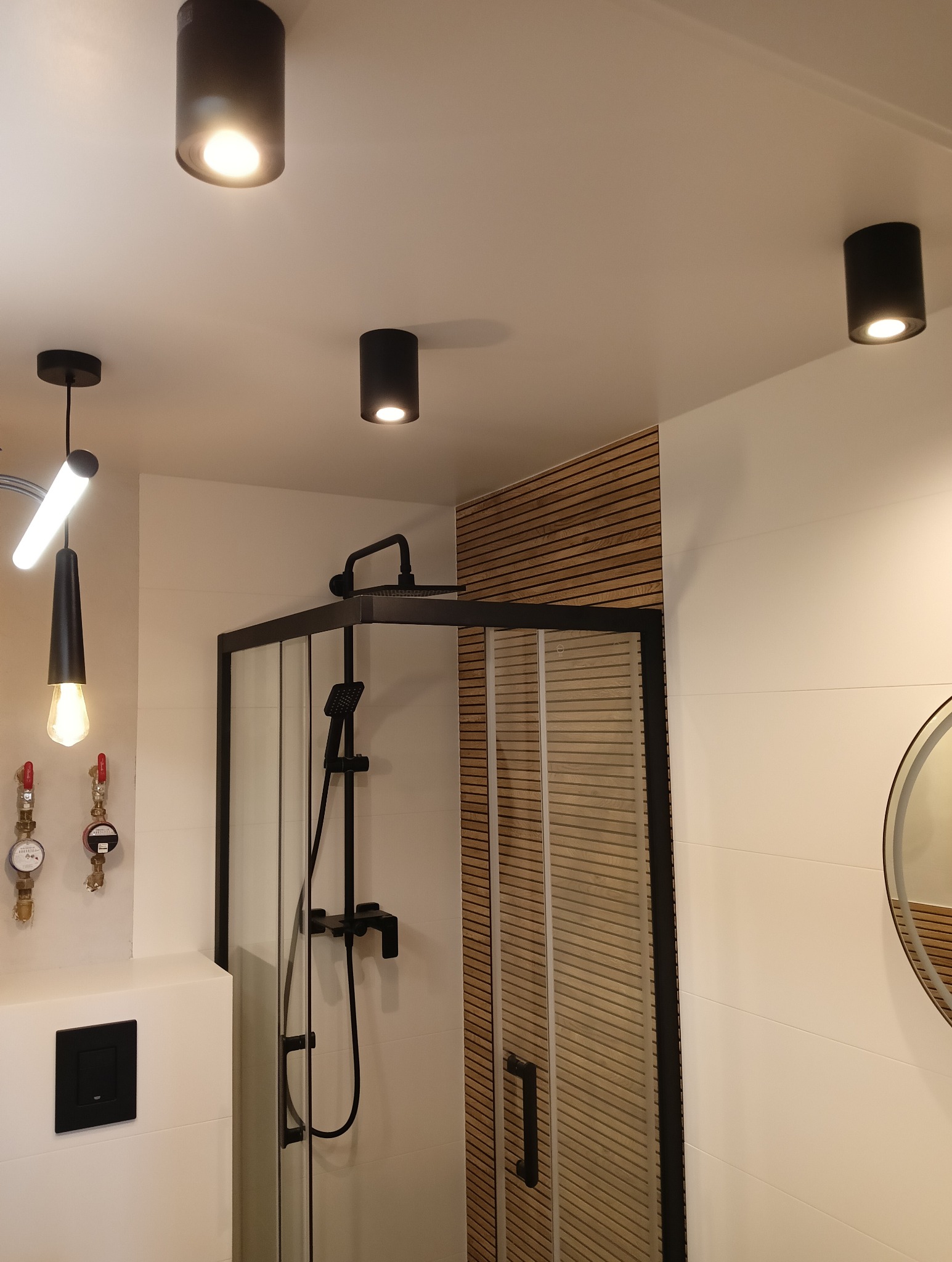 Nowoczesna łazienka z kabiną prysznicową typu walk-in, czarną armaturą i oświetleniem punktowym. Ściana wykończona drewnianymi panelami. Minimalistyczny design.