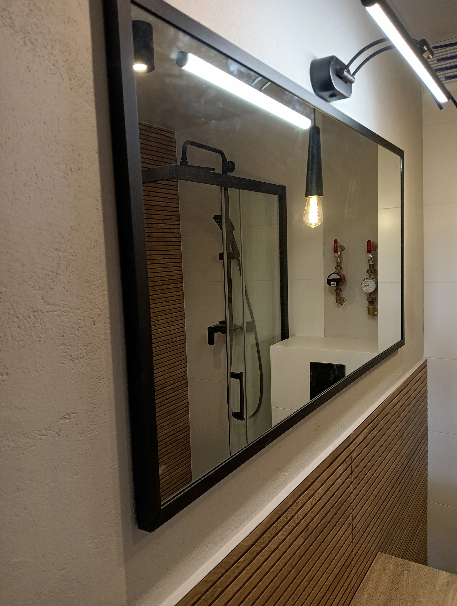 Nowoczesna łazienka z lustrem w czarnej ramie, odbijającym kabinę prysznicową i armaturę. Drewniane panele ścienne dodają ciepła wnętrzu. Oświetlenie punktowe i wisząca lampa.