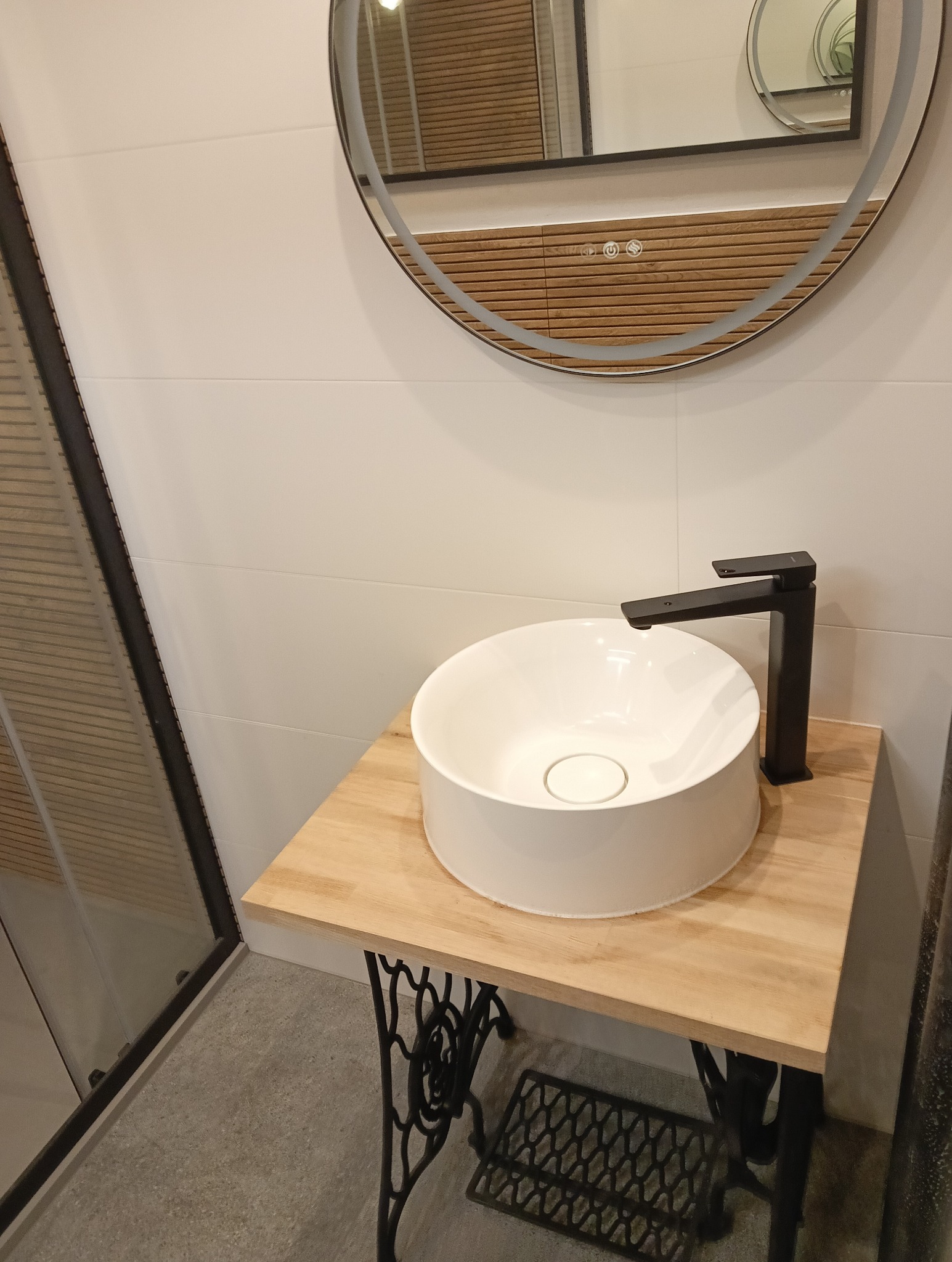 Biała umywalka nablatowa z czarną armaturą na drewnianym blacie wspartym na żeliwnej podstawie maszyny do szycia. W tle białe płytki i okrągłe lustro z podświetleniem.