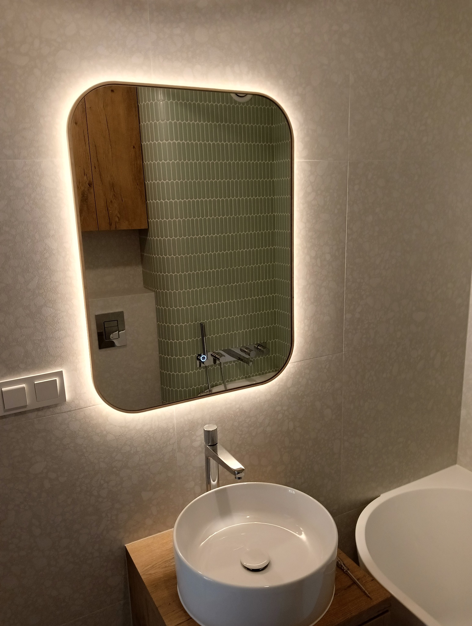 Nowoczesna łazienka z podświetlanym lustrem, umywalką nablatową i zielonymi płytkami w kształcie migdałów odbijającymi się w lustrze.