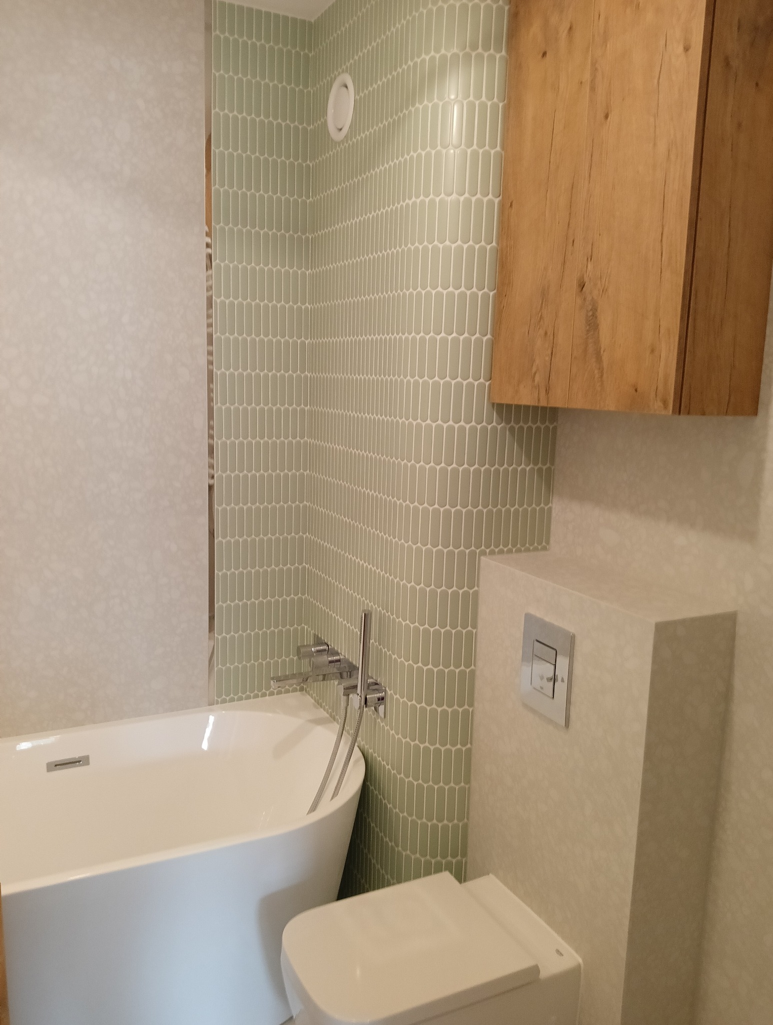 Nowoczesna łazienka z wanną wolnostojącą, toaletą podwieszaną i drewnianą szafką. Ściany wyłożone zieloną mozaiką i jasnymi płytkami z delikatnym wzorem.