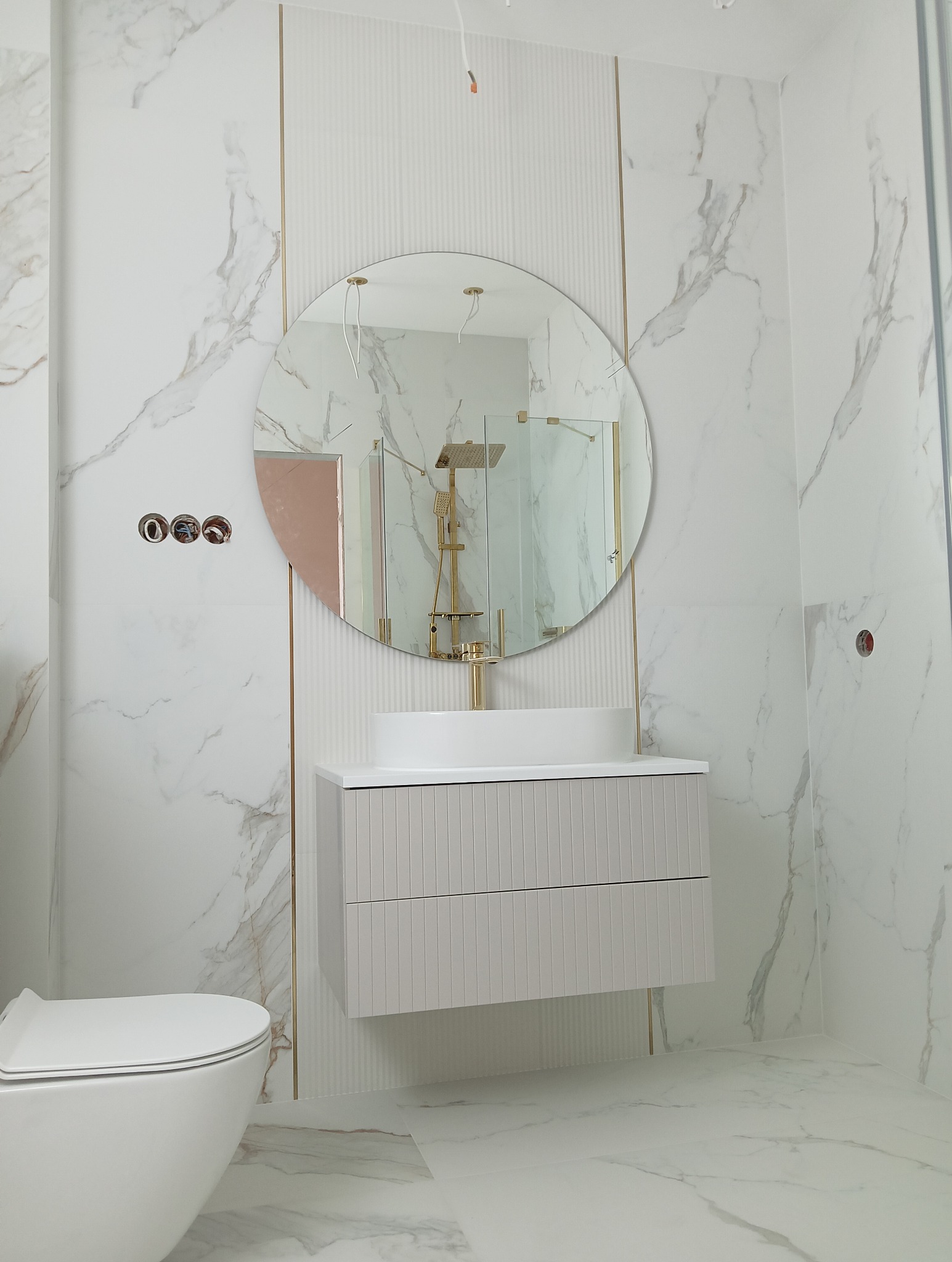 Elegancka łazienka z marmurowymi płytkami, okrągłym lustrem i szafką pod umywalką w odcieniach bieli i szarości. Widoczne złote akcenty i częściowo zamontowana armatura.