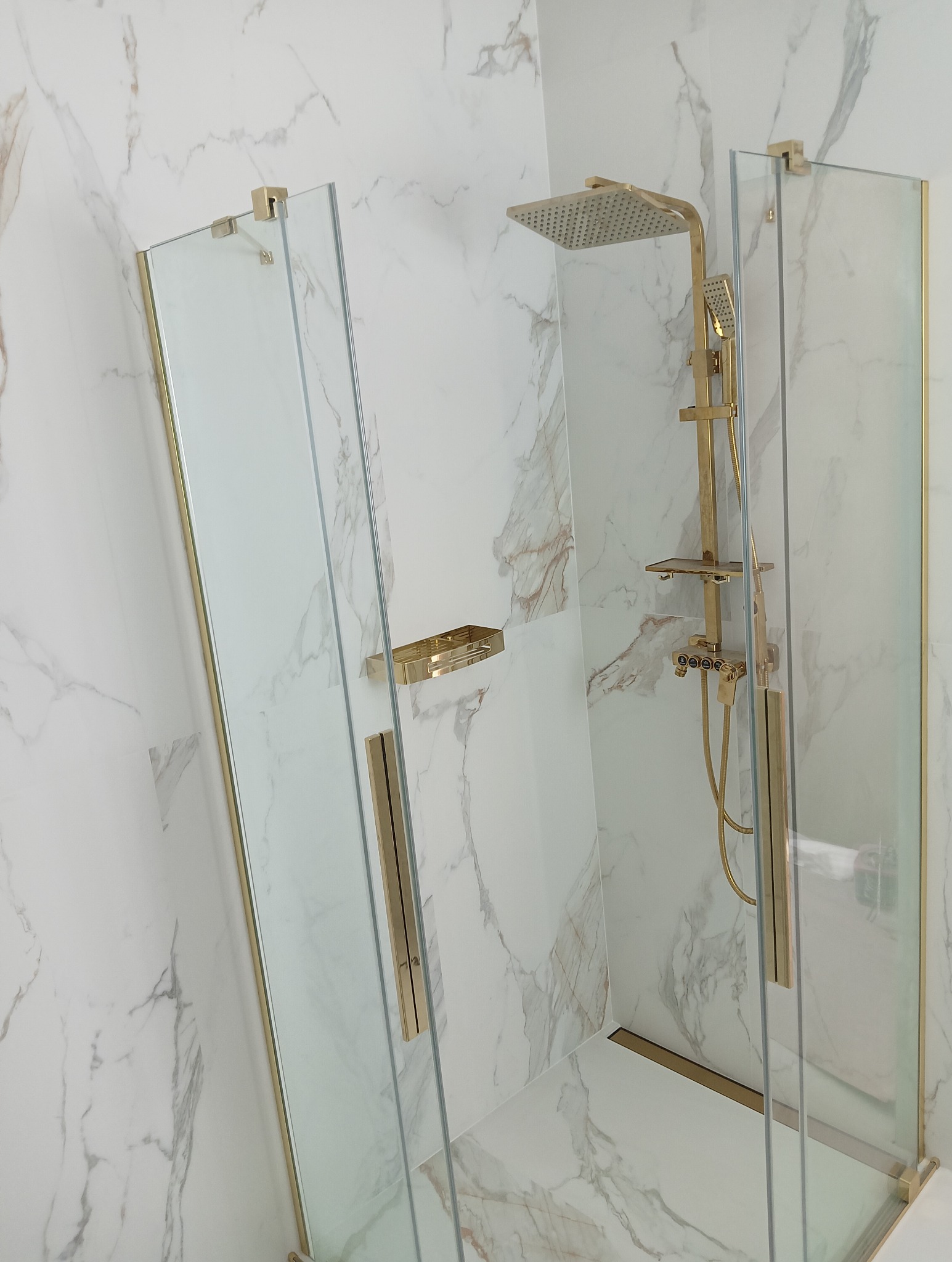 Nowoczesna kabina prysznicowa z złotymi elementami na tle marmurowych płytek. Szklane ścianki kabiny z eleganckimi uchwytami i deszczownicą.