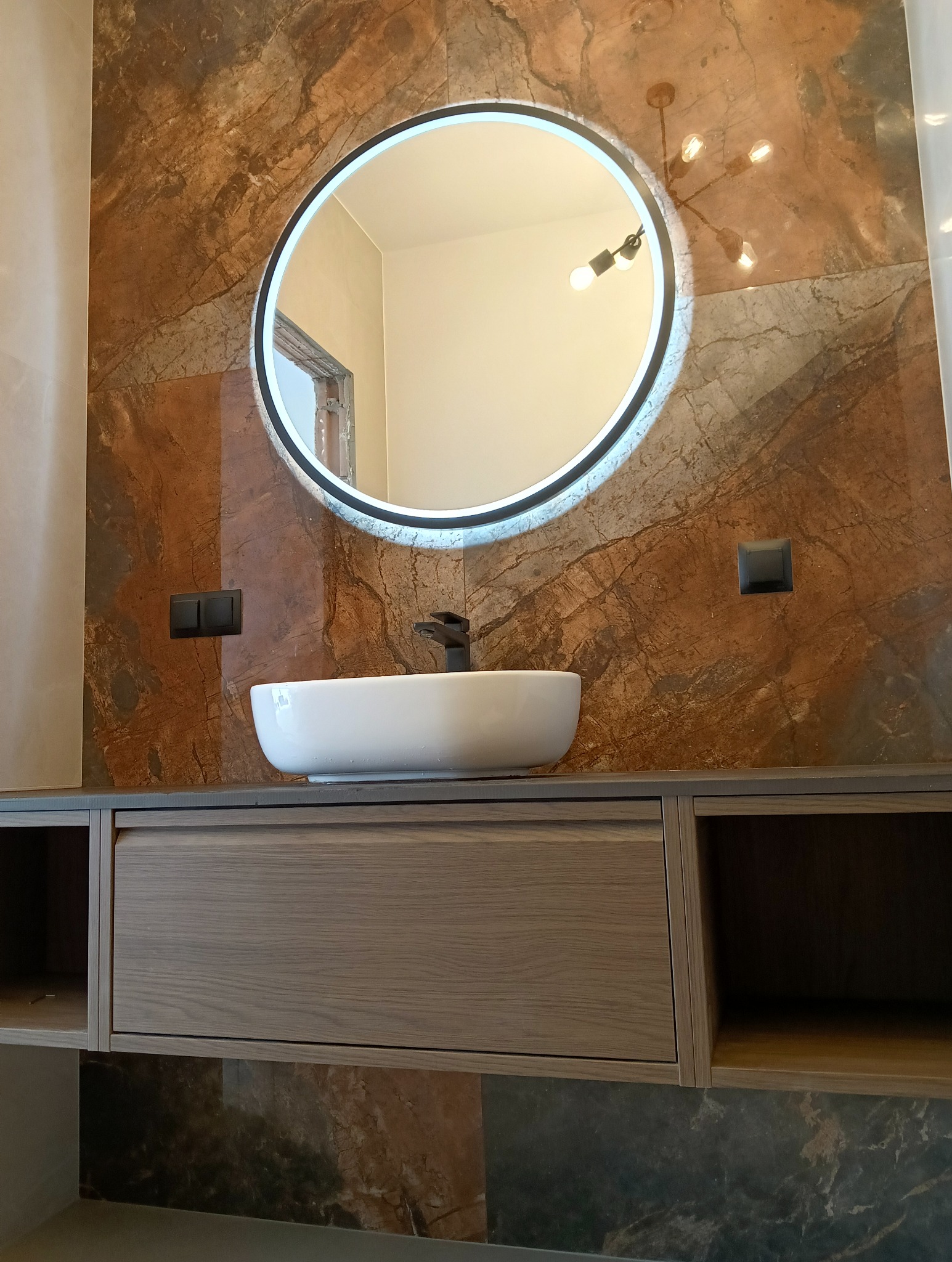 Nowoczesna łazienka z okrągłym lustrem LED, umywalką nablatową i szafką z drewnianym frontem na tle ściany z płytek imitujących marmur w odcieniach brązu i beżu.