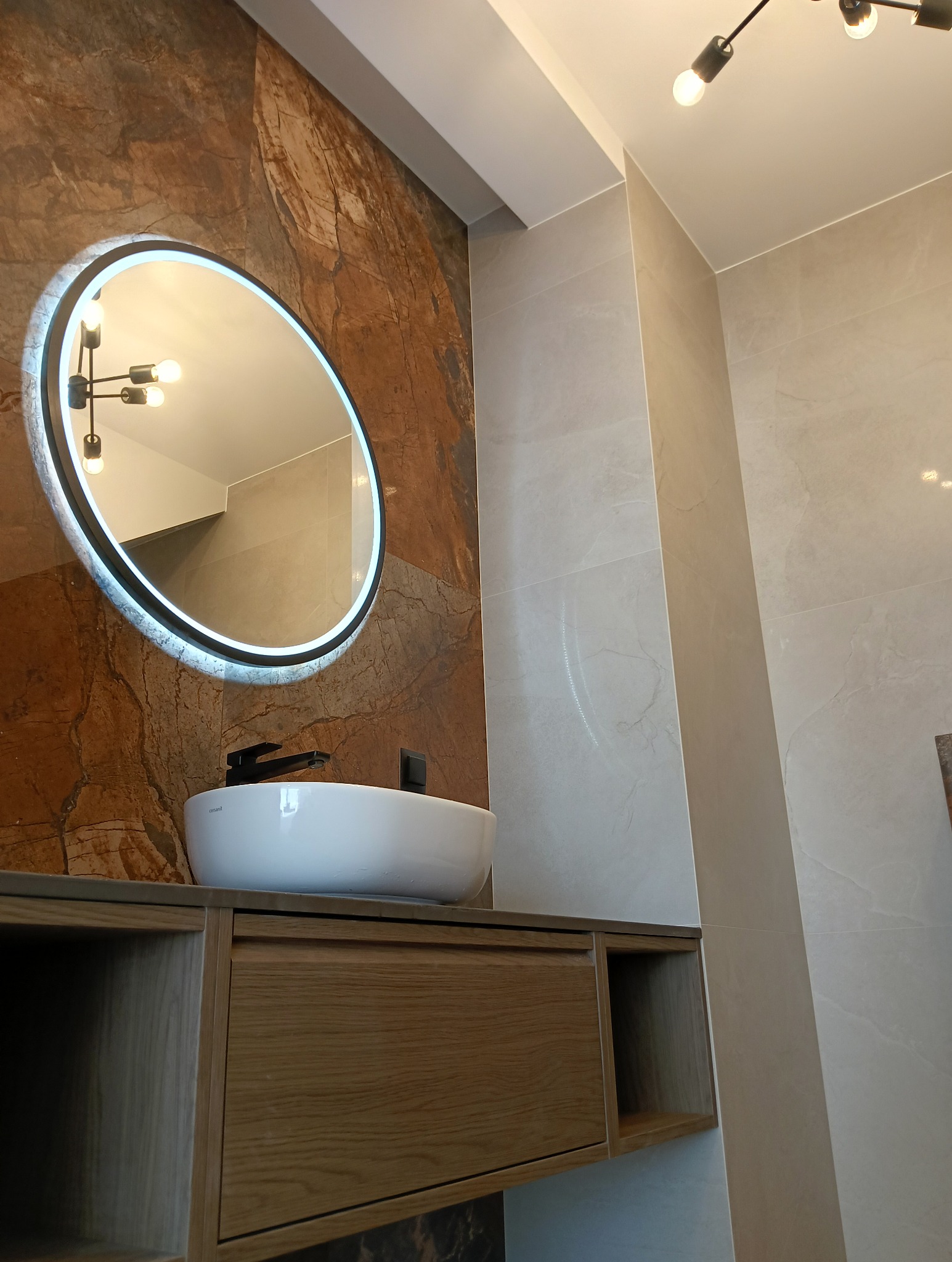 Nowoczesna łazienka: okrągłe lustro LED nad białą umywalką nablatową z czarną armaturą, drewniana szafka pod umywalką, płytki imitujące kamień na ścianach.