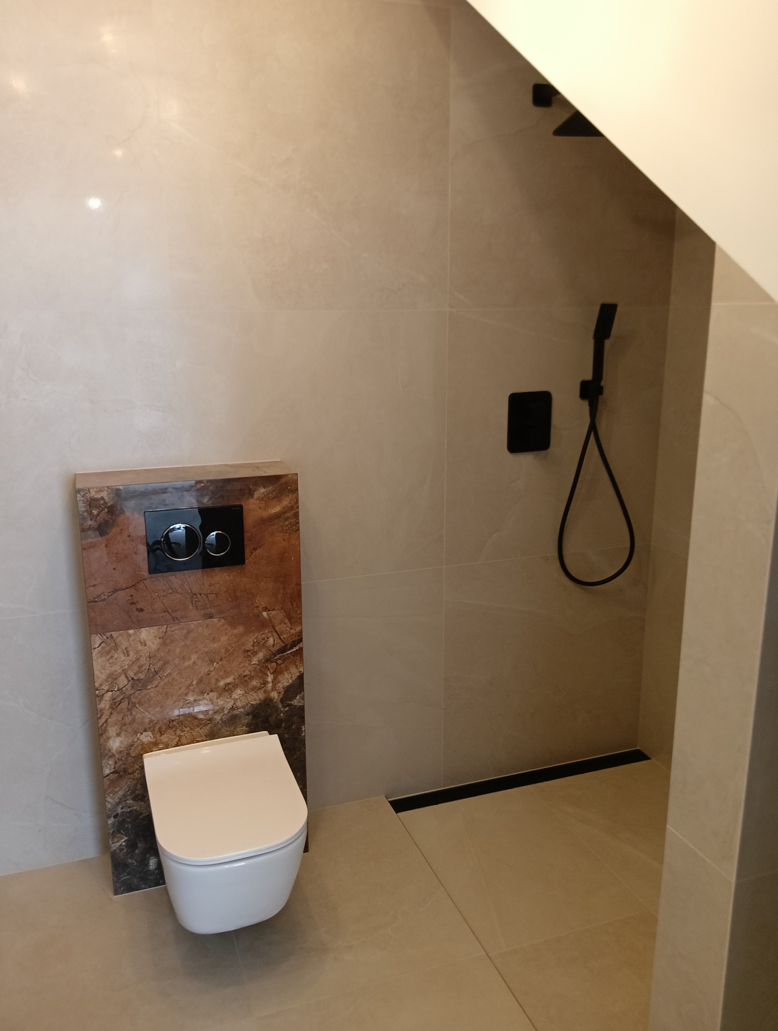 Nowoczesna łazienka z beżowymi płytkami, podwieszaną toaletą na tle marmurowej obudowy i czarną armaturą prysznicową. Minimalistyczny design, eleganckie wykończenie.