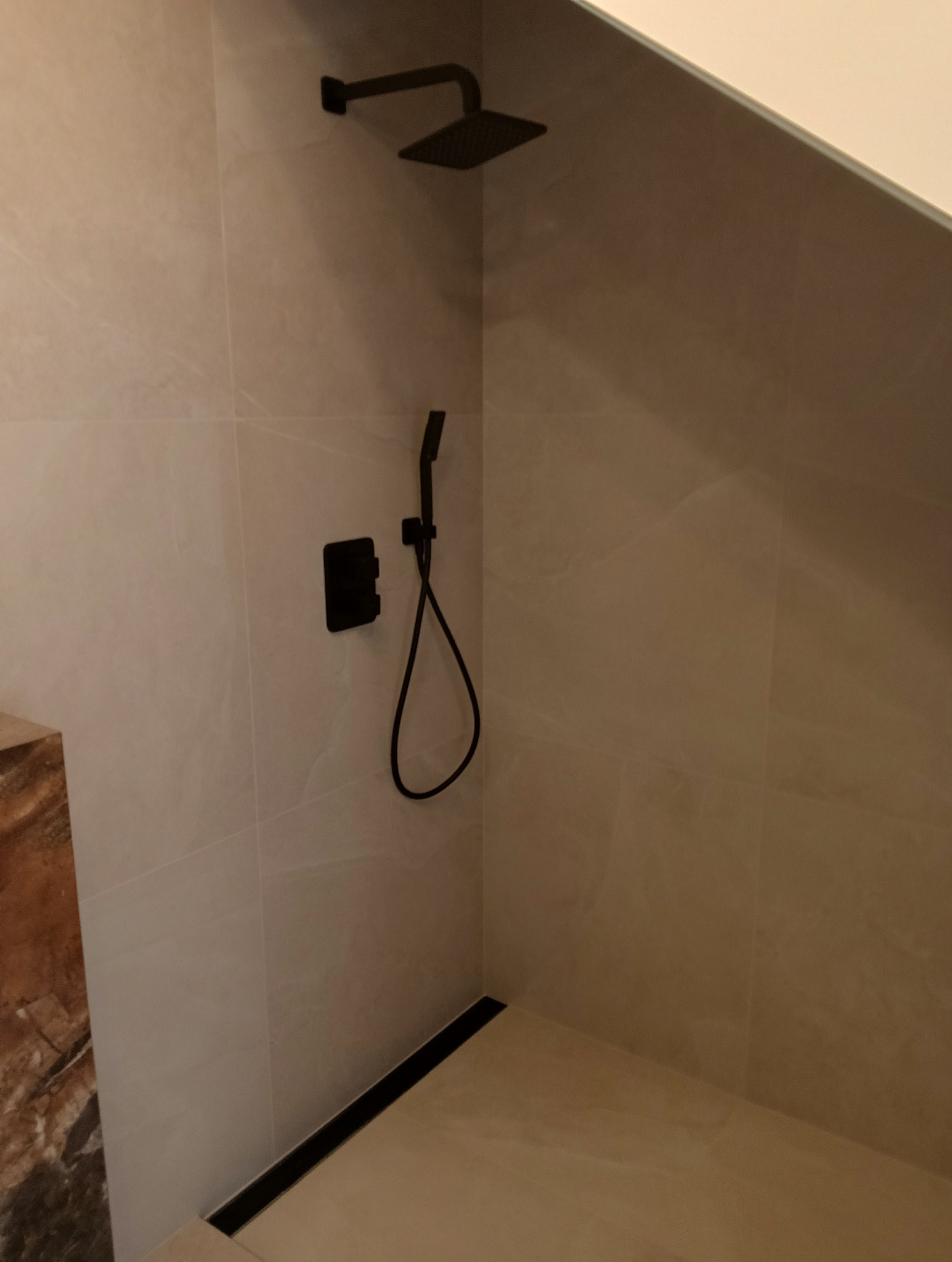 Nowoczesna kabina prysznicowa walk-in z czarną armaturą i odpływem liniowym. Beżowe płytki imitujące marmur na ścianach i podłodze. Minimalistyczny design.